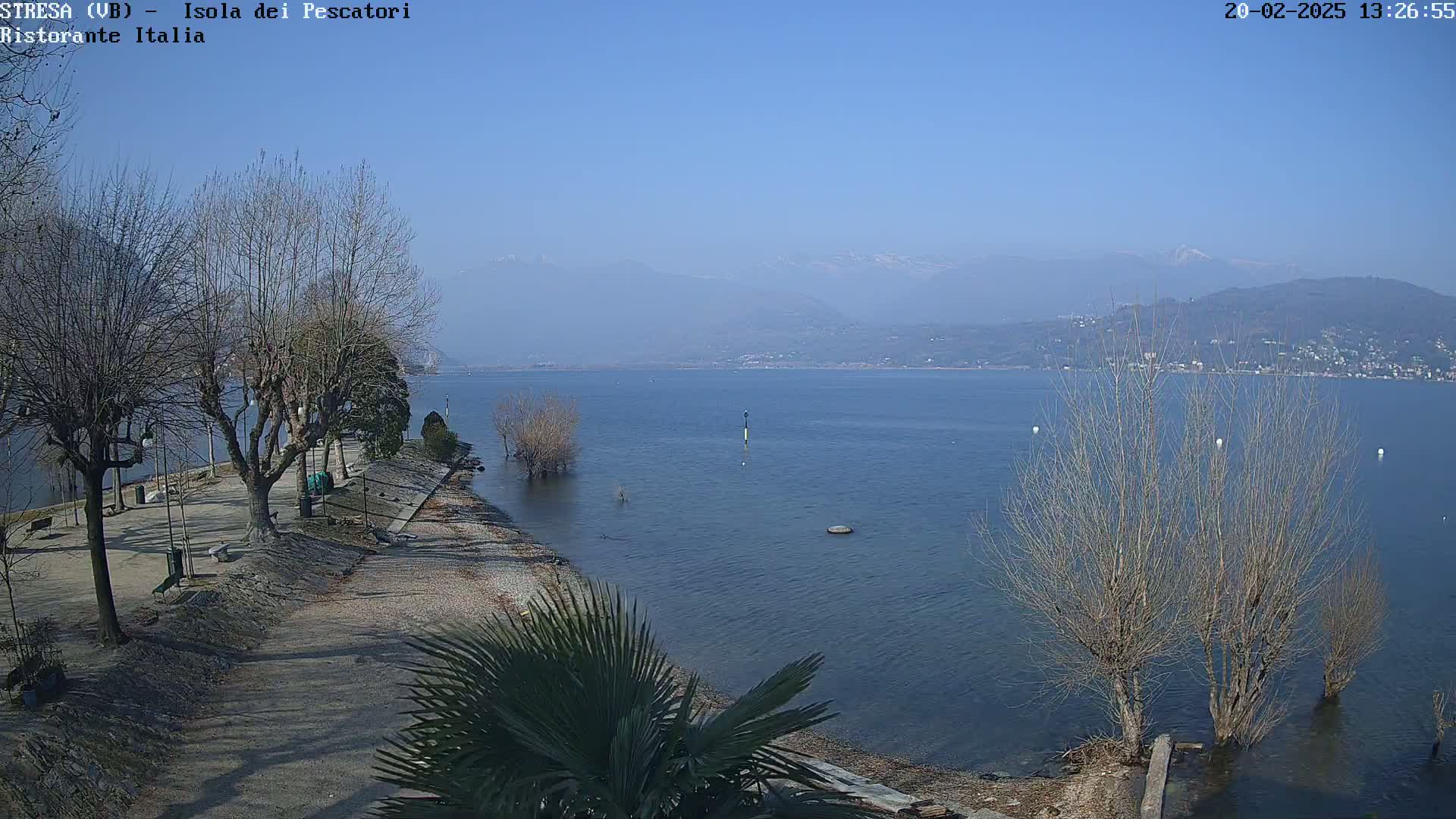 A clear, sunny day overlooks a calm lake with bare trees lining a shoreline path, and mountains in the background.