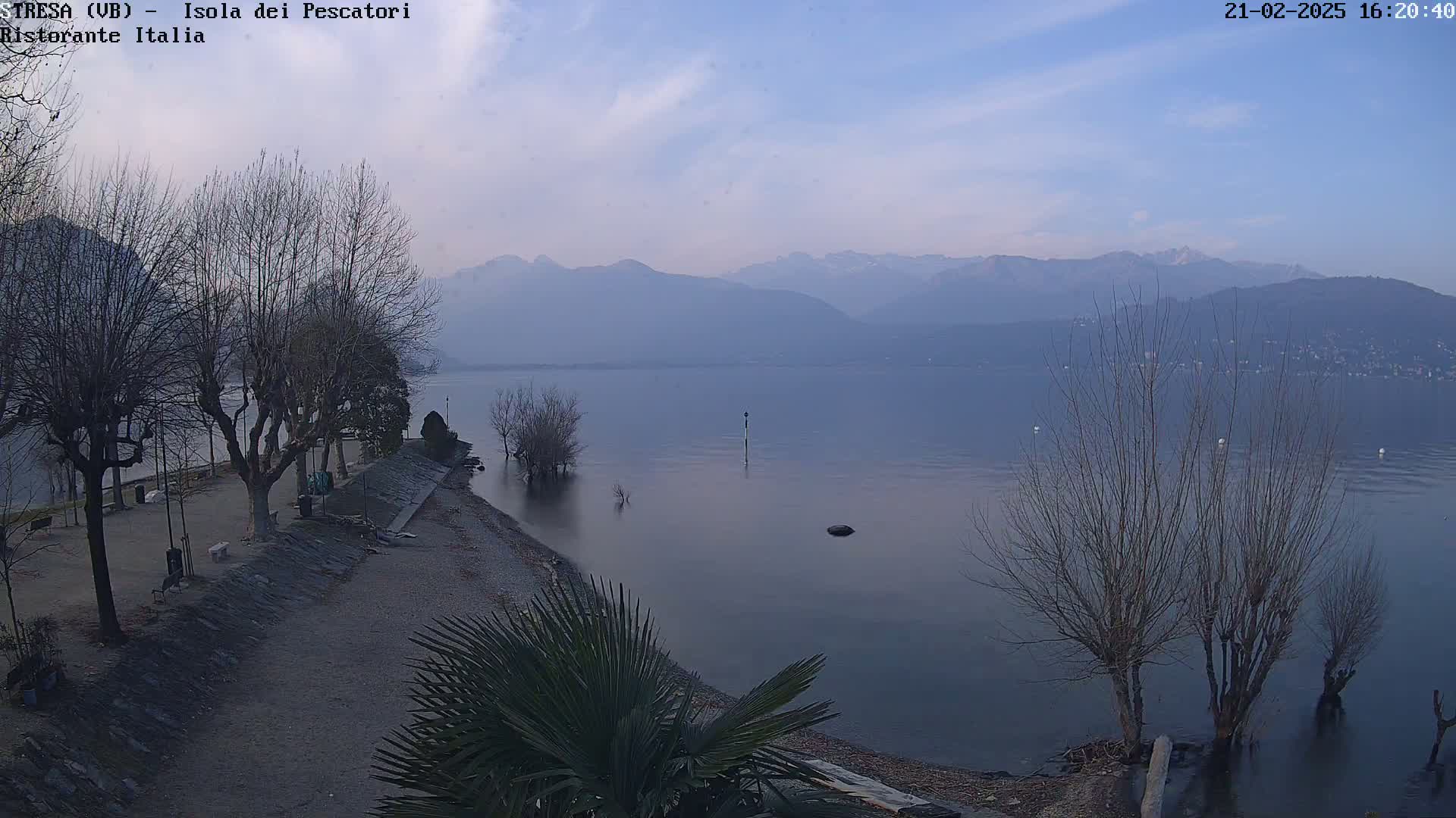 A calm lake stretches toward hazy mountains under a pale blue sky, with bare trees lining a shoreline path.