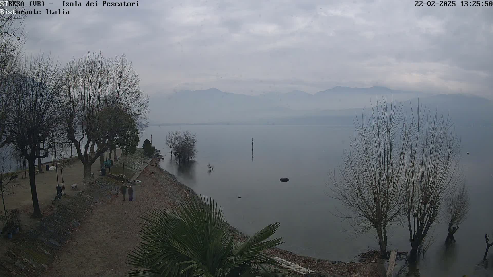 A calm lake with hazy mountains in the background under an overcast sky, a shoreline with bare trees and a few people walking.