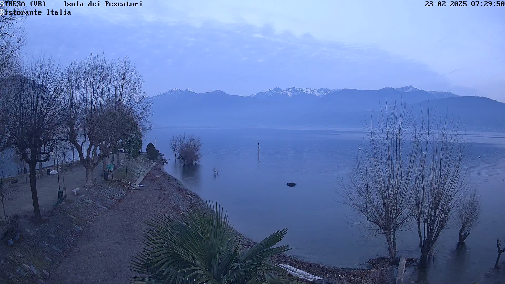 A calm, blue lake under a pale sky stretches toward snow-capped mountains in the distance, with leafless trees lining a gravel shoreline.