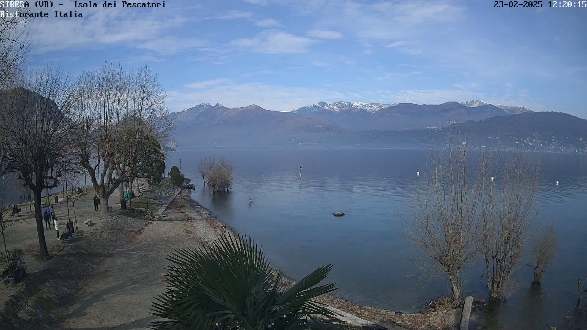A calm lake with snow-capped mountains in the background, under a mostly clear blue sky, is seen from a lakeside park with bare trees and a few people walking.