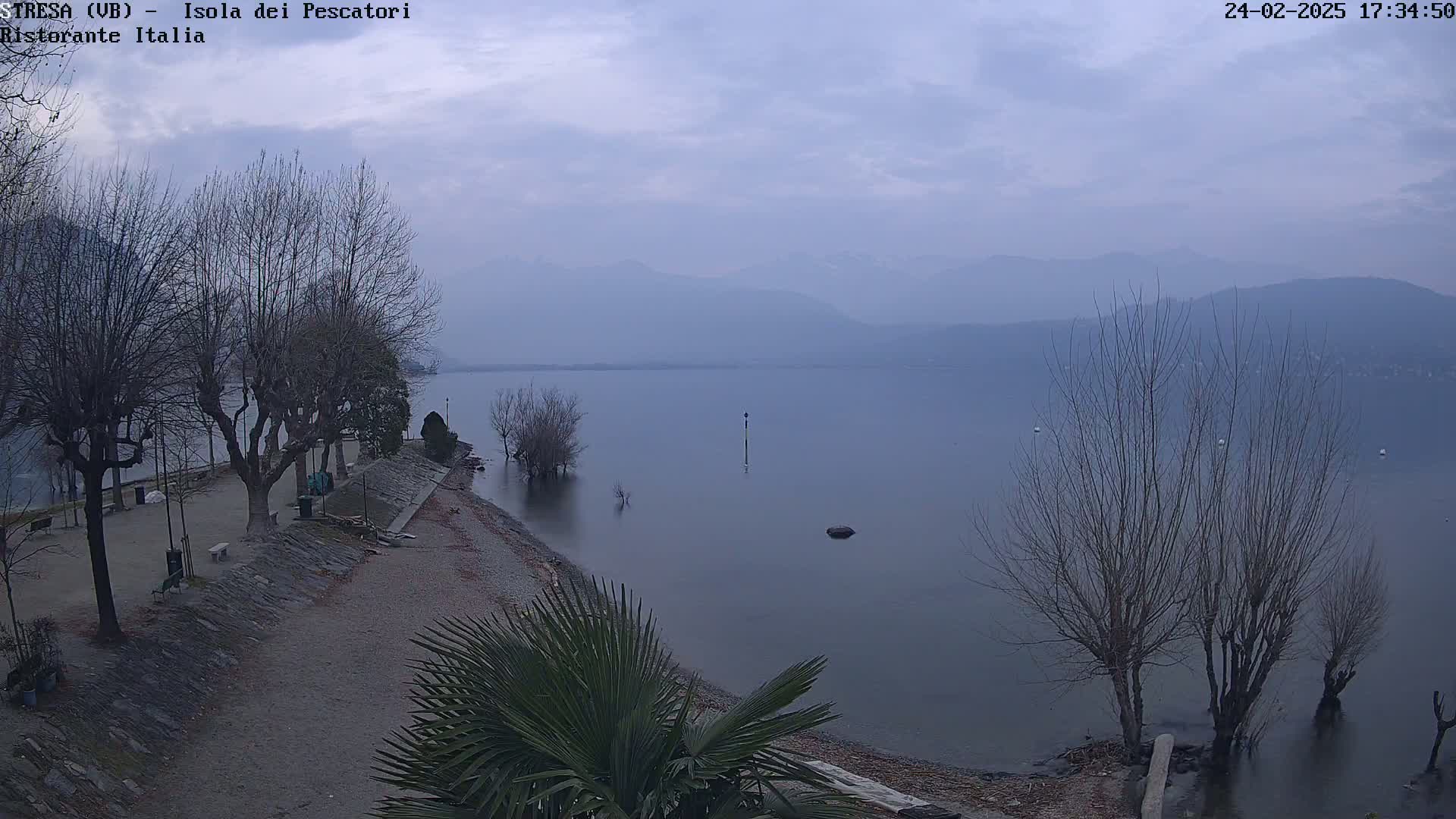 A tranquil lake scene under an overcast sky shows bare trees lining a pebble shoreline with a calm body of water extending to hazy mountains in the distance.