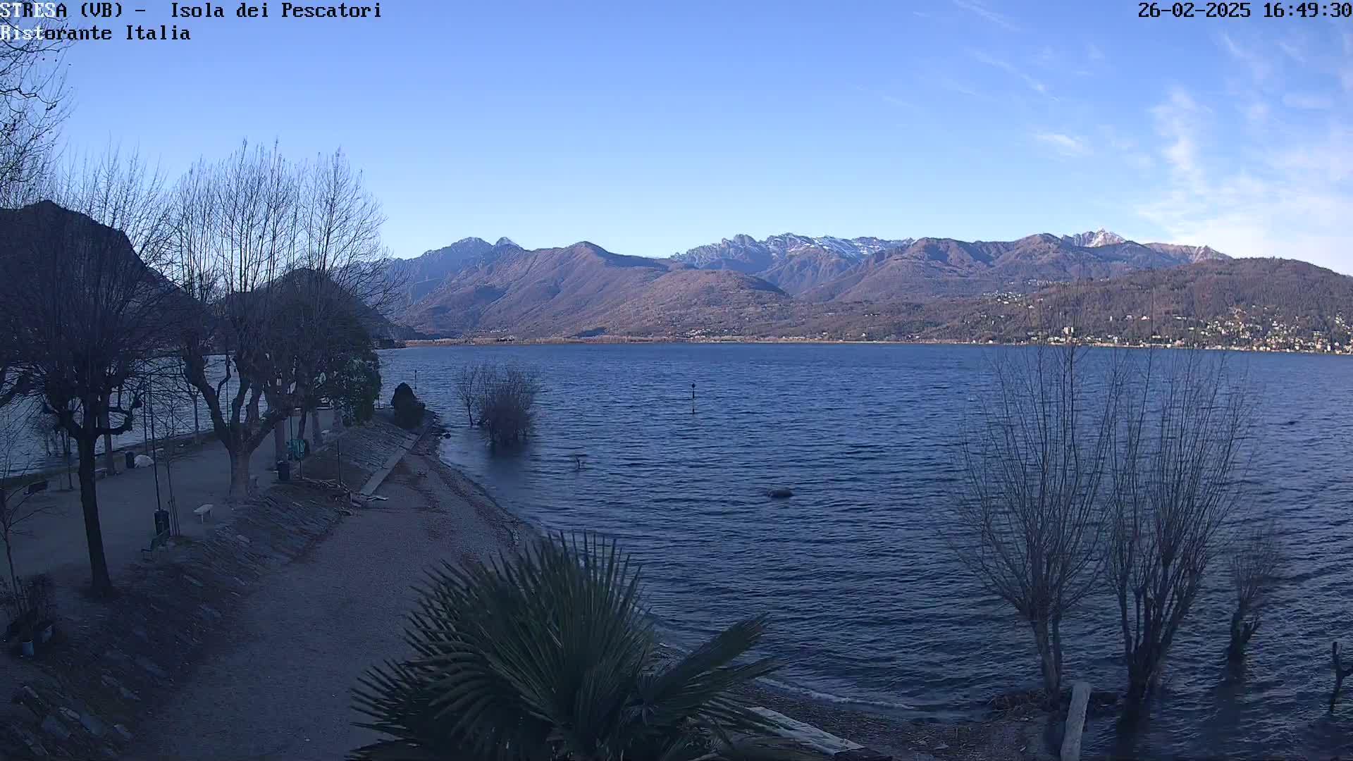 A calm lake is nestled between snow-capped mountains under a clear blue sky, with bare trees lining a sandy shoreline.