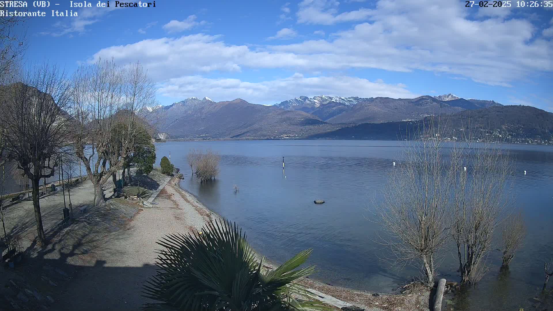 A partly sunny day shows a calm lake, snow-capped mountains in the background, and a shoreline with bare trees and a gravel beach.
