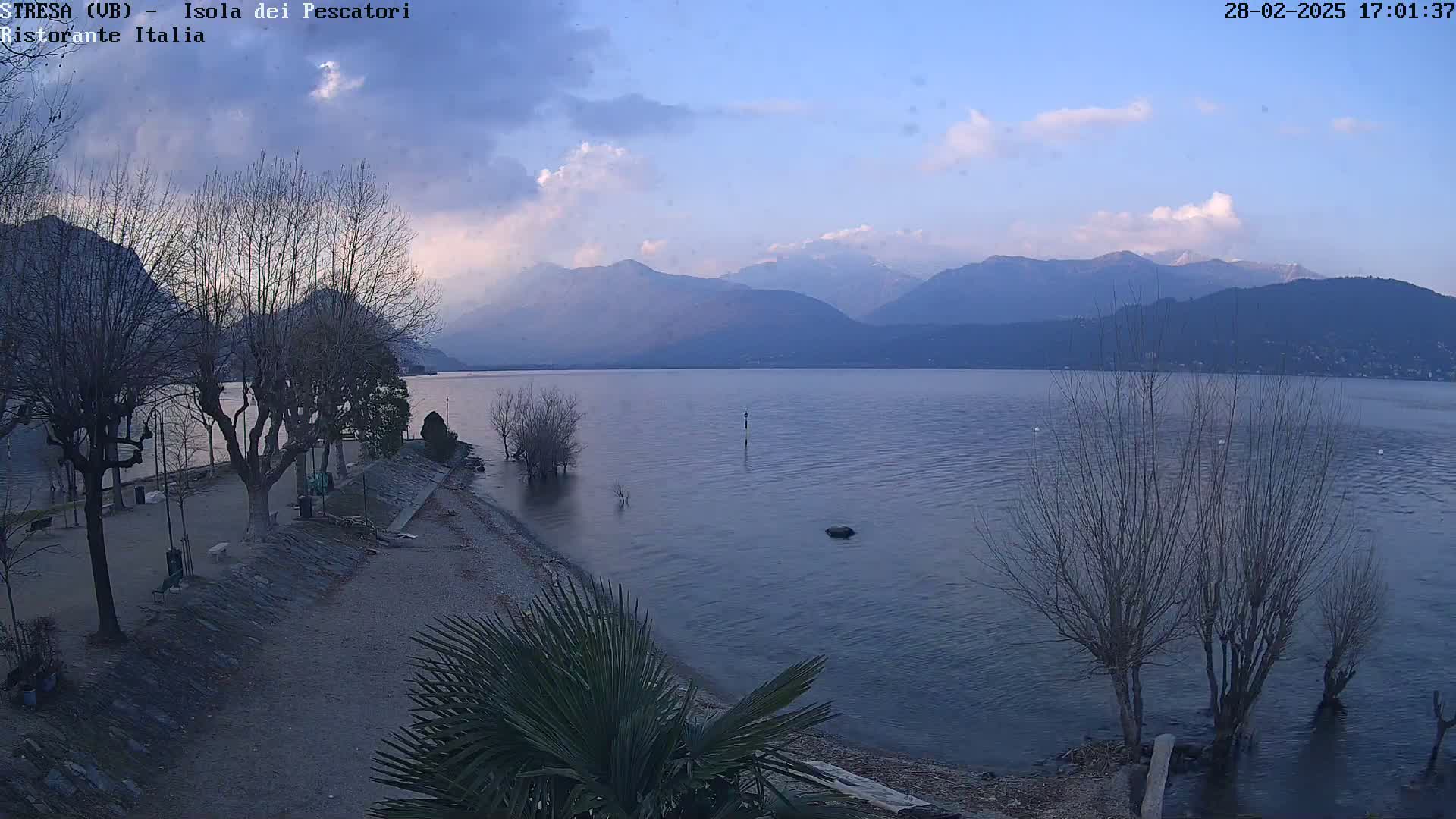 A tranquil lake scene under a partly cloudy sky shows bare trees lining a calm shoreline with mountains in the background.