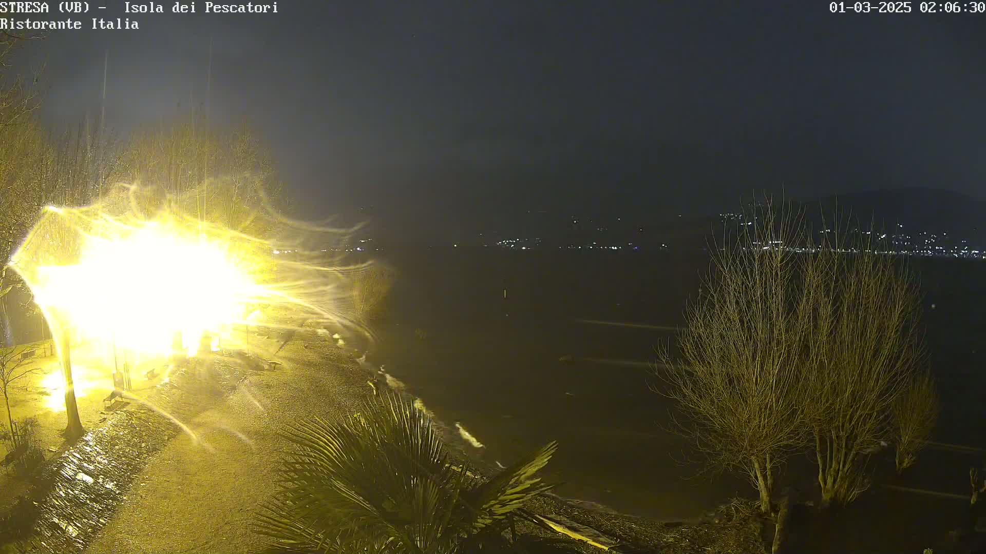 A bright light illuminates a snowy or windy scene at night near a body of water, with distant lights visible across the water.