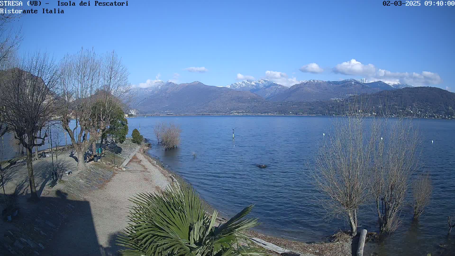 A mostly sunny day reveals a calm lake shore with leafless trees, a small beach, and snow-capped mountains in the distance.