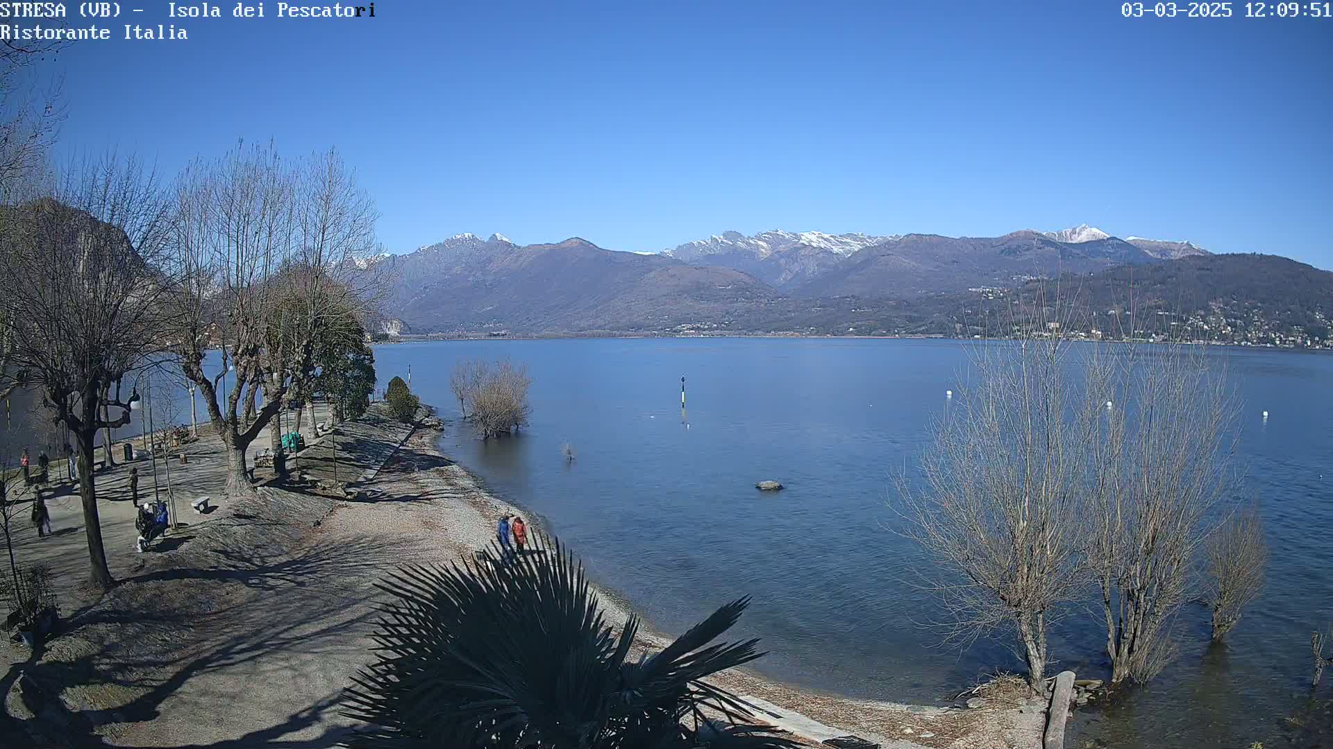 A clear, sunny day reveals a calm lake with snow-capped mountains in the background, a shoreline with bare trees and a few people, and a sandy beach.