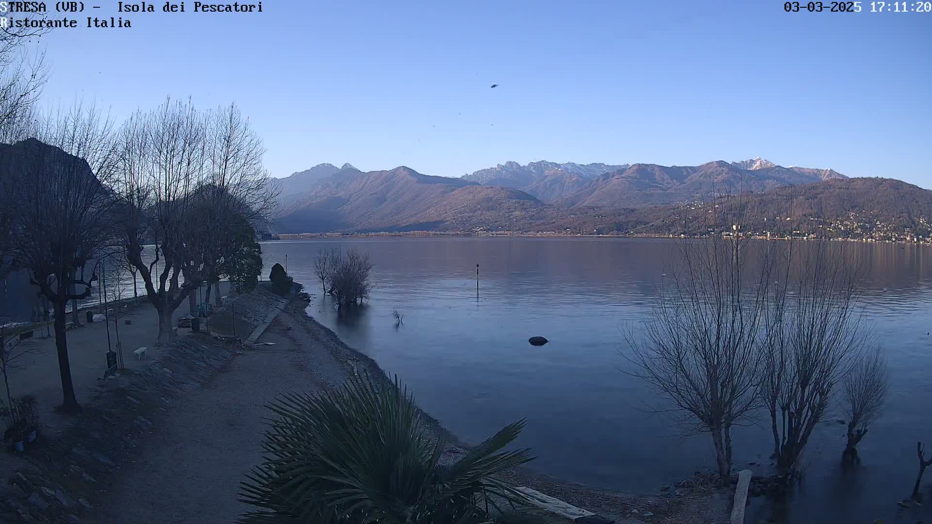 A calm lake stretches out before a shoreline with bare trees and a mountain range in the background under a clear, light blue sky.