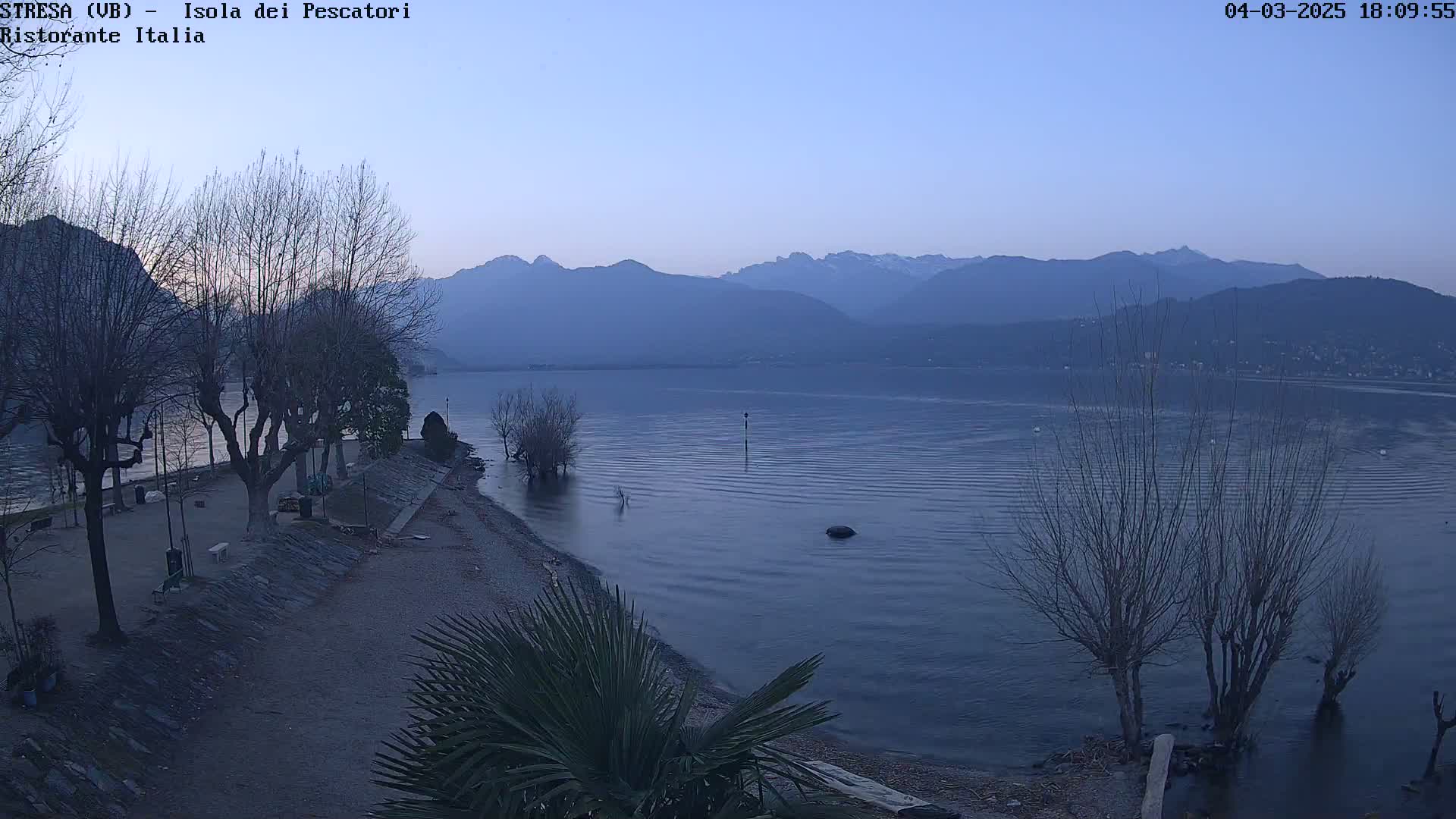A calm lake at dusk, under a clear sky, is bordered by a shoreline with bare trees and a small sandy area, with mountains visible in the distance.