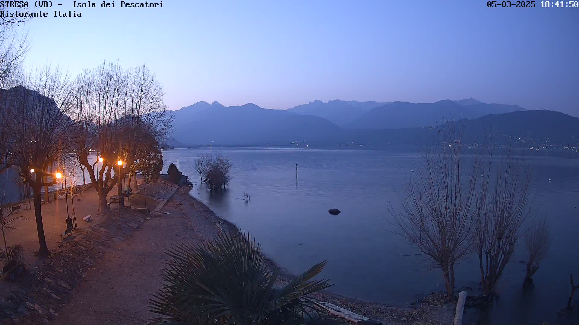 A calm lake at dusk is seen from a shoreline with bare trees and a few lights, backed by distant mountains under a twilight sky.