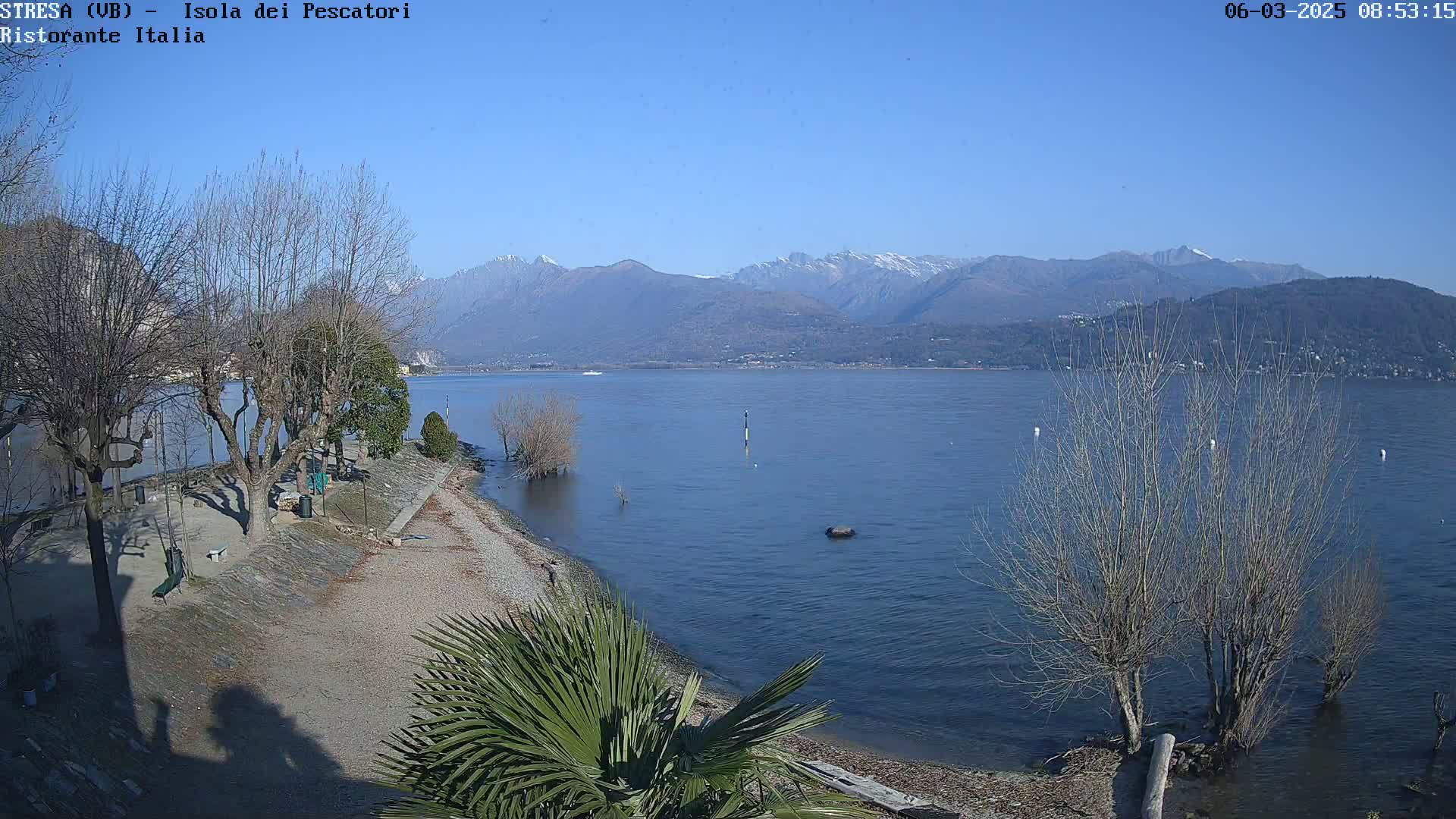 A tranquil lake shore with leafless trees and a small sandy beach, under a clear blue sky with snow-capped mountains in the distance.
