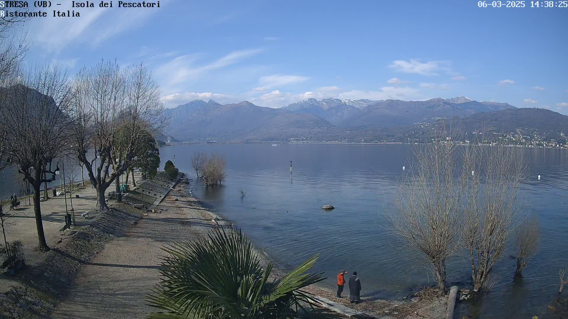 Two people stand on a lakeshore path near bare trees, overlooking a calm lake with snow-capped mountains in the background under a mostly clear sky.
