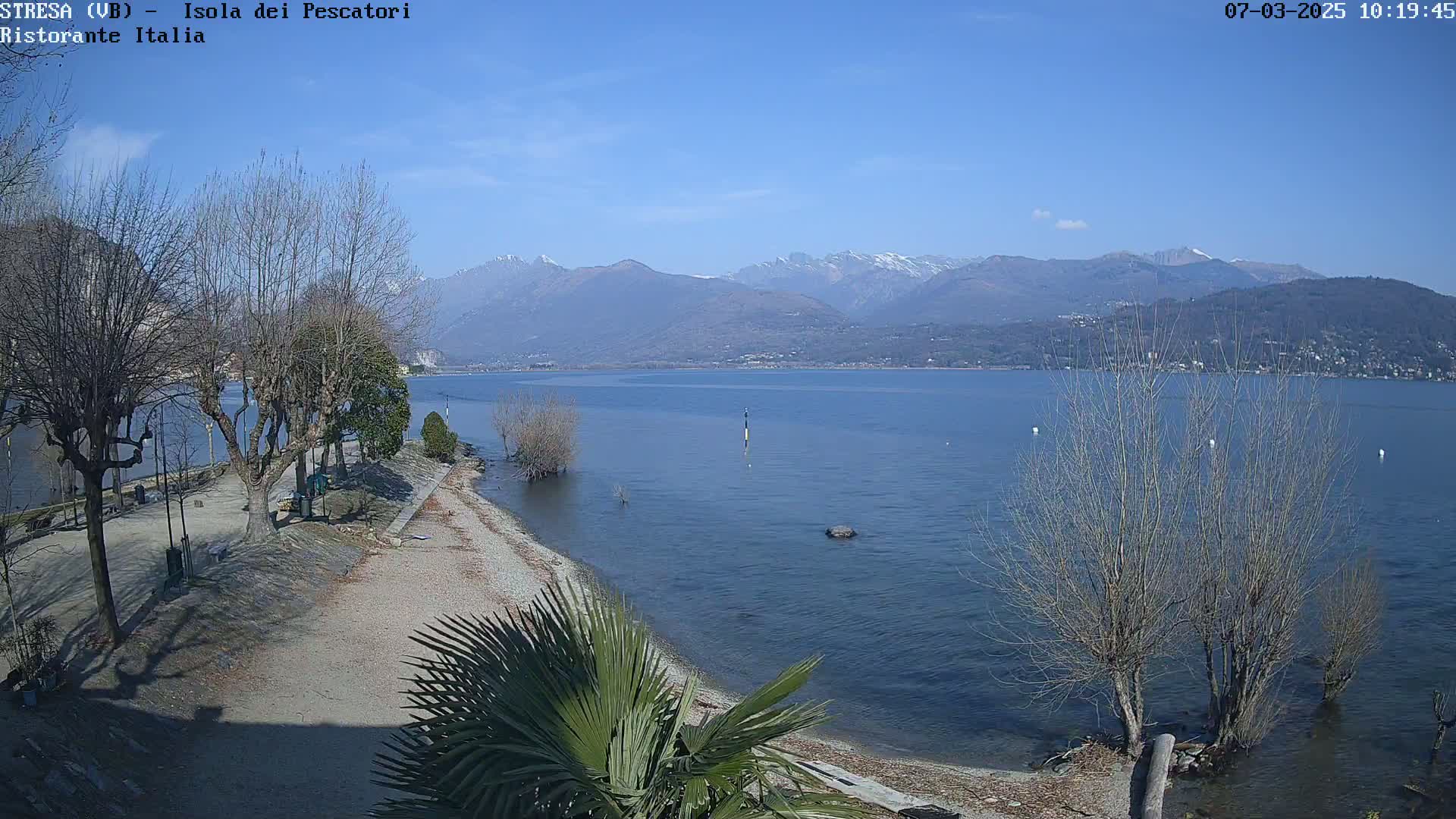 A clear, sunny day shows a calm lake with a pebble beach and leafless trees in the foreground, mountains in the background.