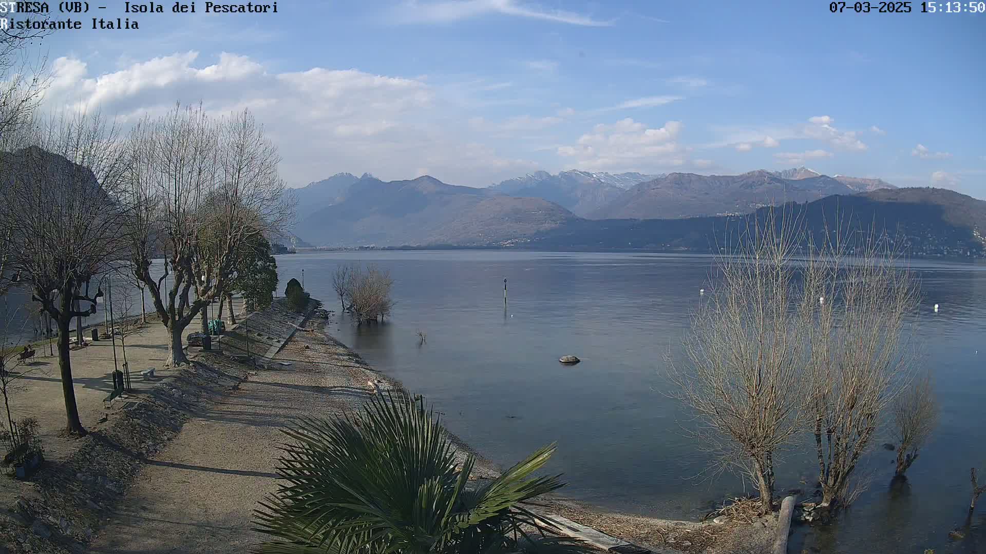A calm lake with mountains in the background, under a partly cloudy sky, is seen from a lakeside park with bare trees and a few palm trees.