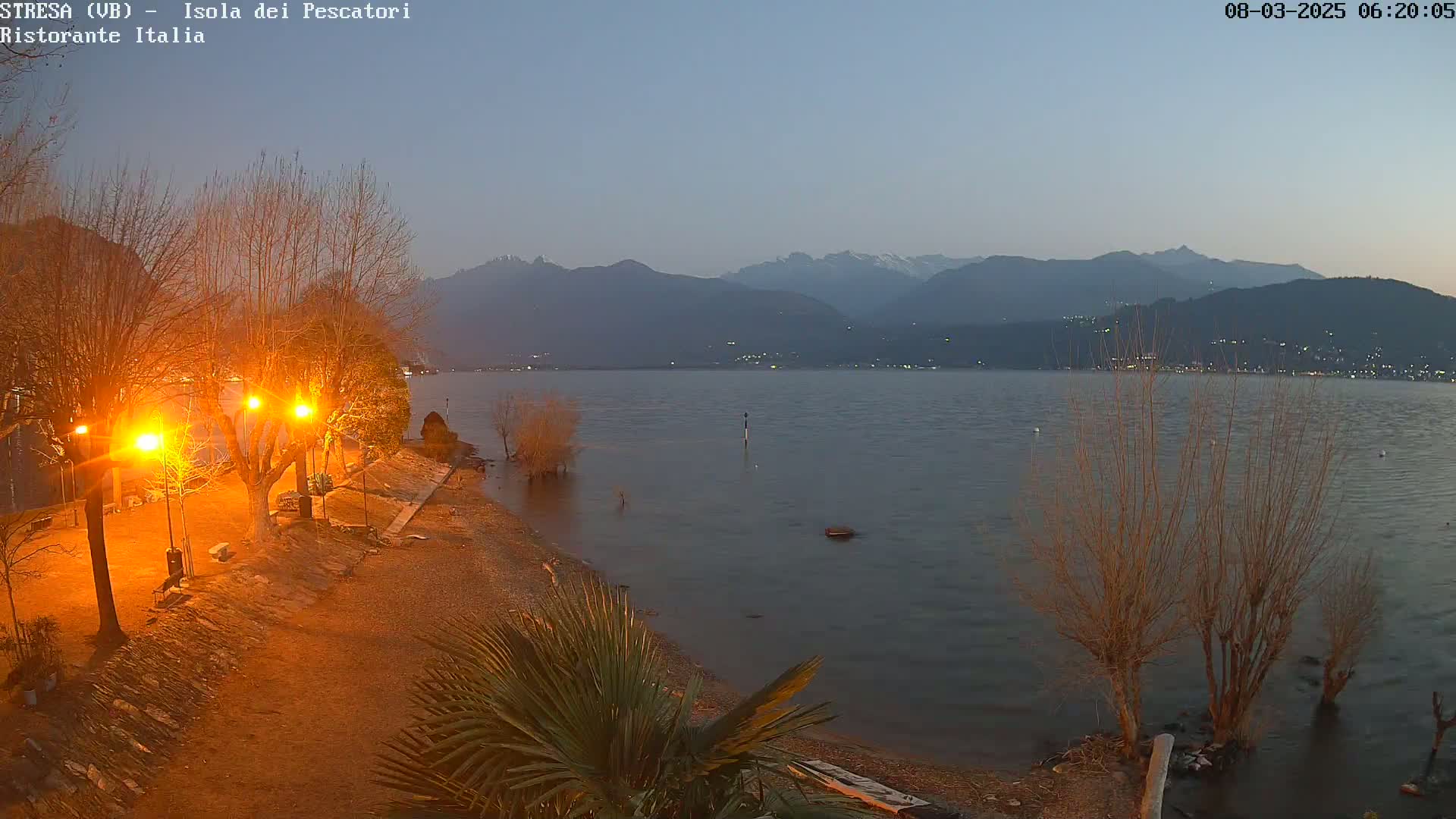 A dimly lit lakeshore at twilight shows bare trees illuminated by orange streetlights, a calm lake extending to snow-capped mountains in the distance.