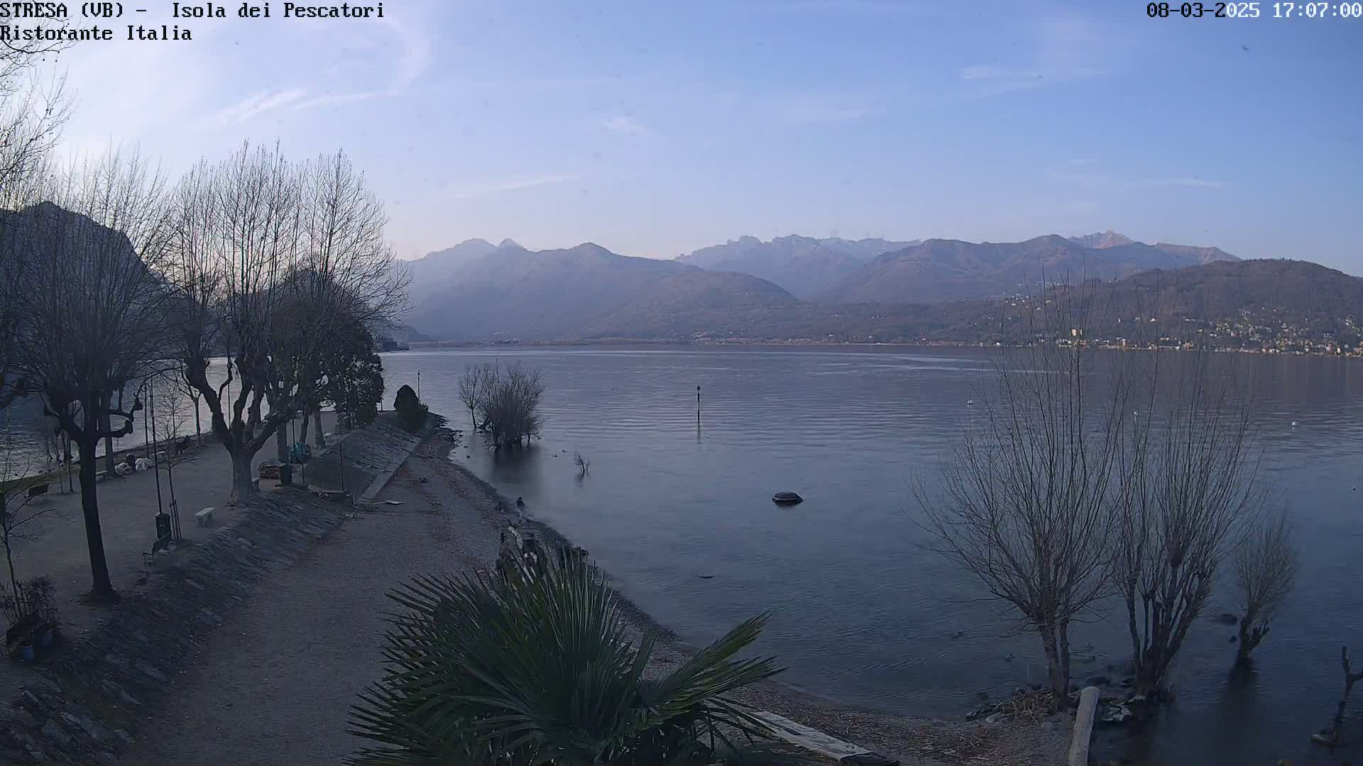 A calm lake under a clear, pale blue sky is backed by distant mountains, with bare trees lining a shoreline path.