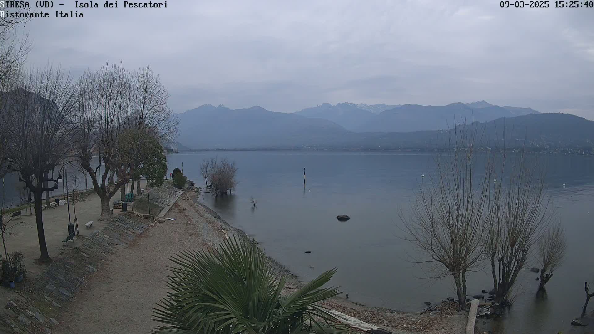 A calm lake with bare trees lining a pebble shore, mountains in the background under an overcast sky.
