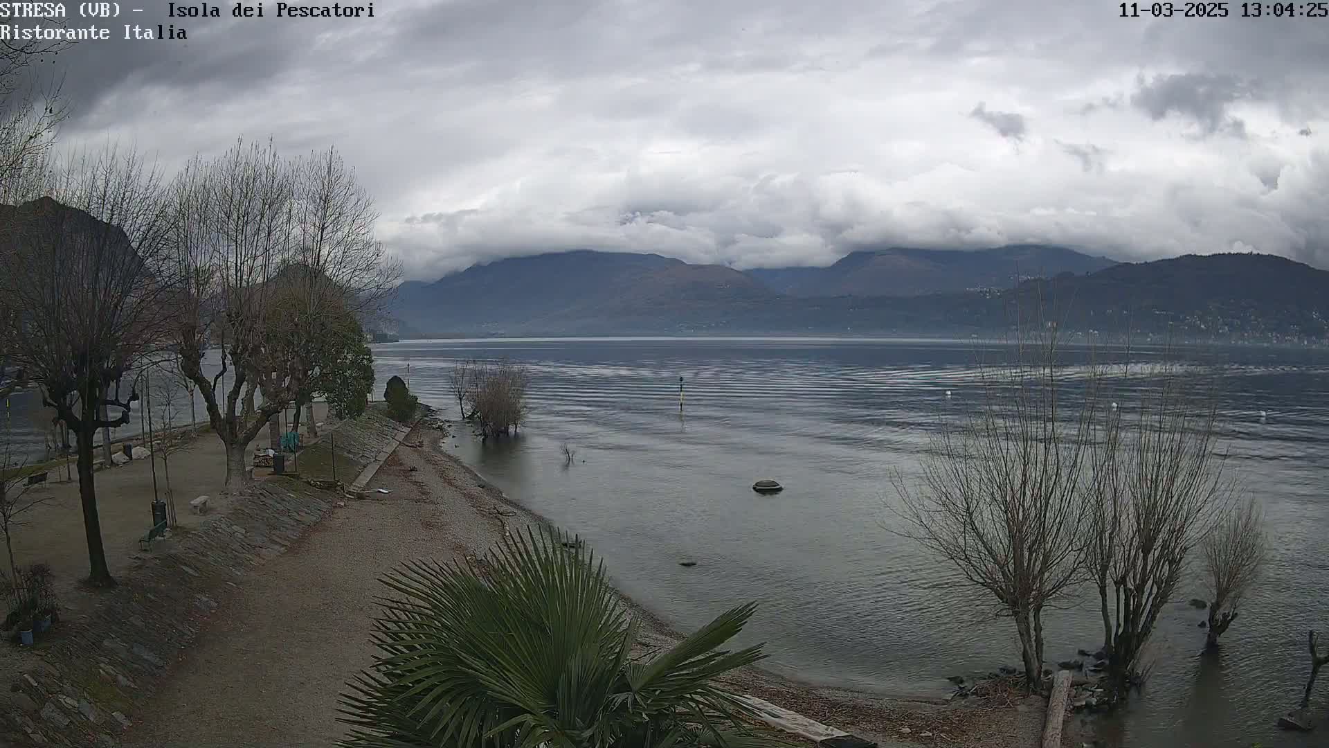 A mostly cloudy day reveals a calm lake with mountains in the background, a shoreline with leafless trees and a paved area with benches.