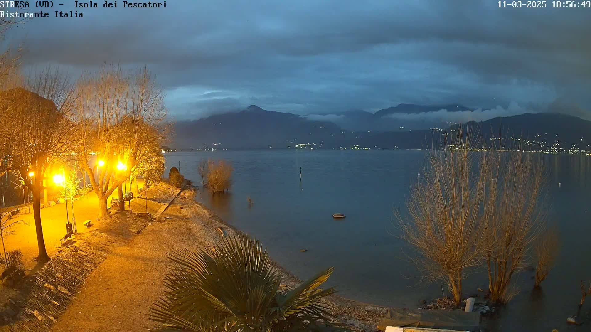 A calm lake at twilight is seen from a shoreline with illuminated trees and a sandy beach, mountains faintly visible in the background under a mostly cloudy sky.