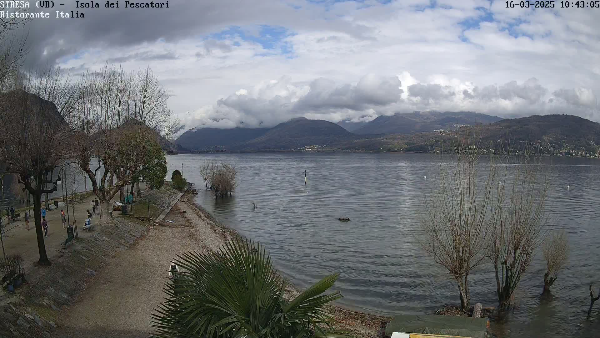 A lake with a shoreline featuring bare trees, a sandy beach, and some people near buildings under a partly cloudy sky, backed by mountains.
