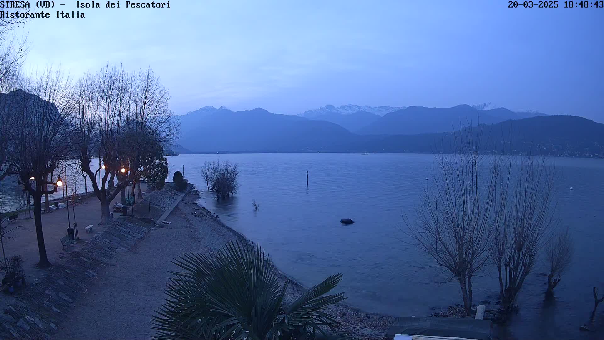 A tranquil lakeside scene at dusk shows a calm body of water reflecting the silhouettes of distant mountains under a clear, twilight sky.
