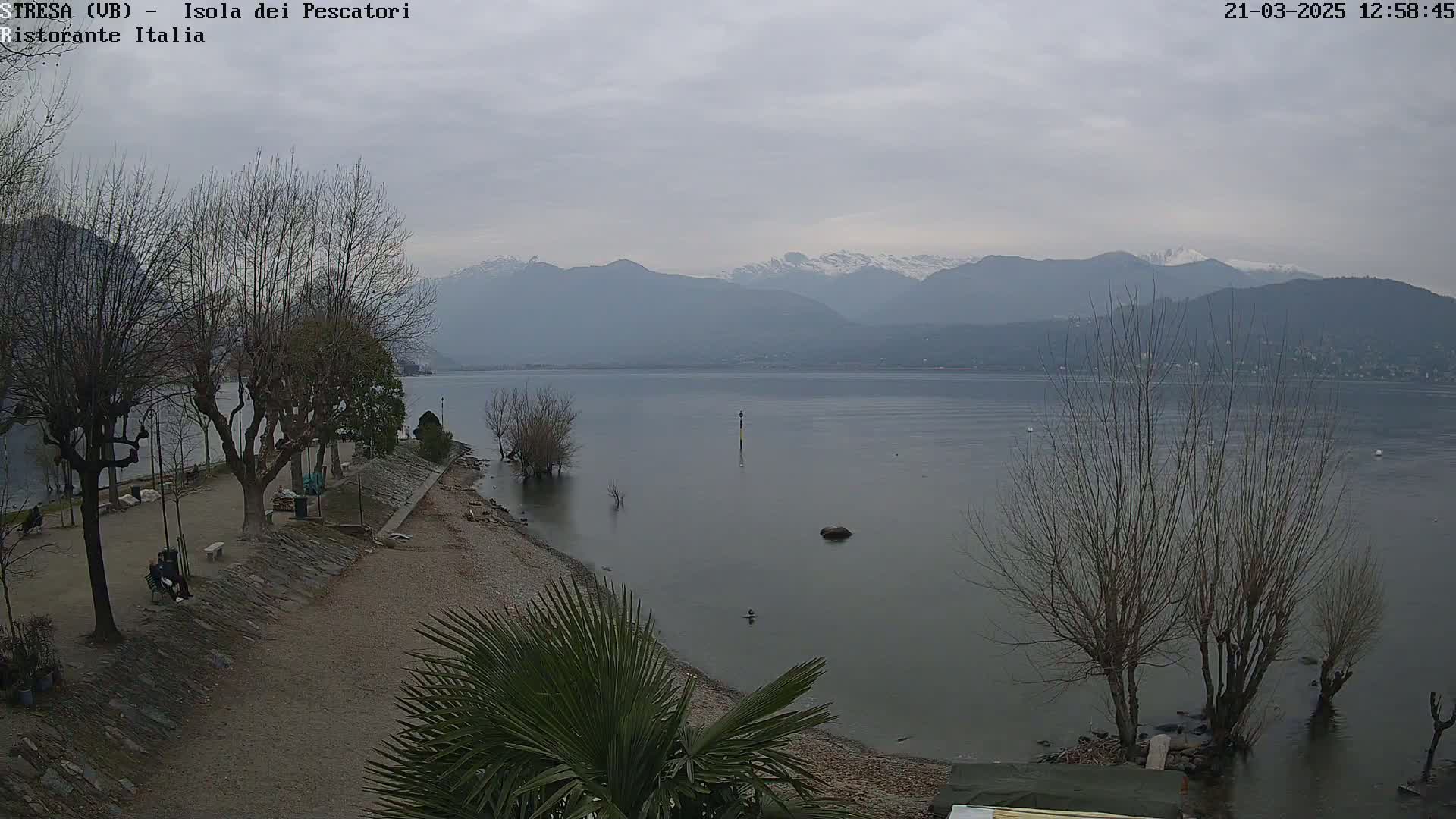 A calm lake stretches toward snow-capped mountains under an overcast sky, with bare trees lining a shoreline featuring a pebble beach and a paved walkway.