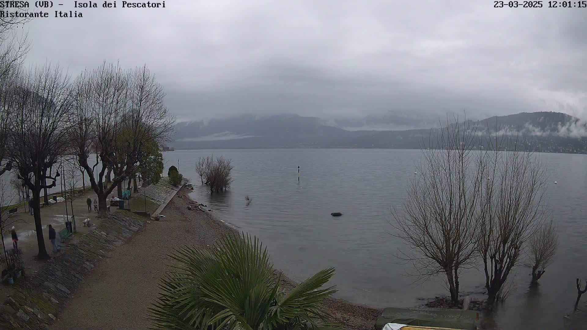 A gray, overcast day reveals a calm lake with bare trees lining a shoreline path where several people walk; mountains are visible in the distance.