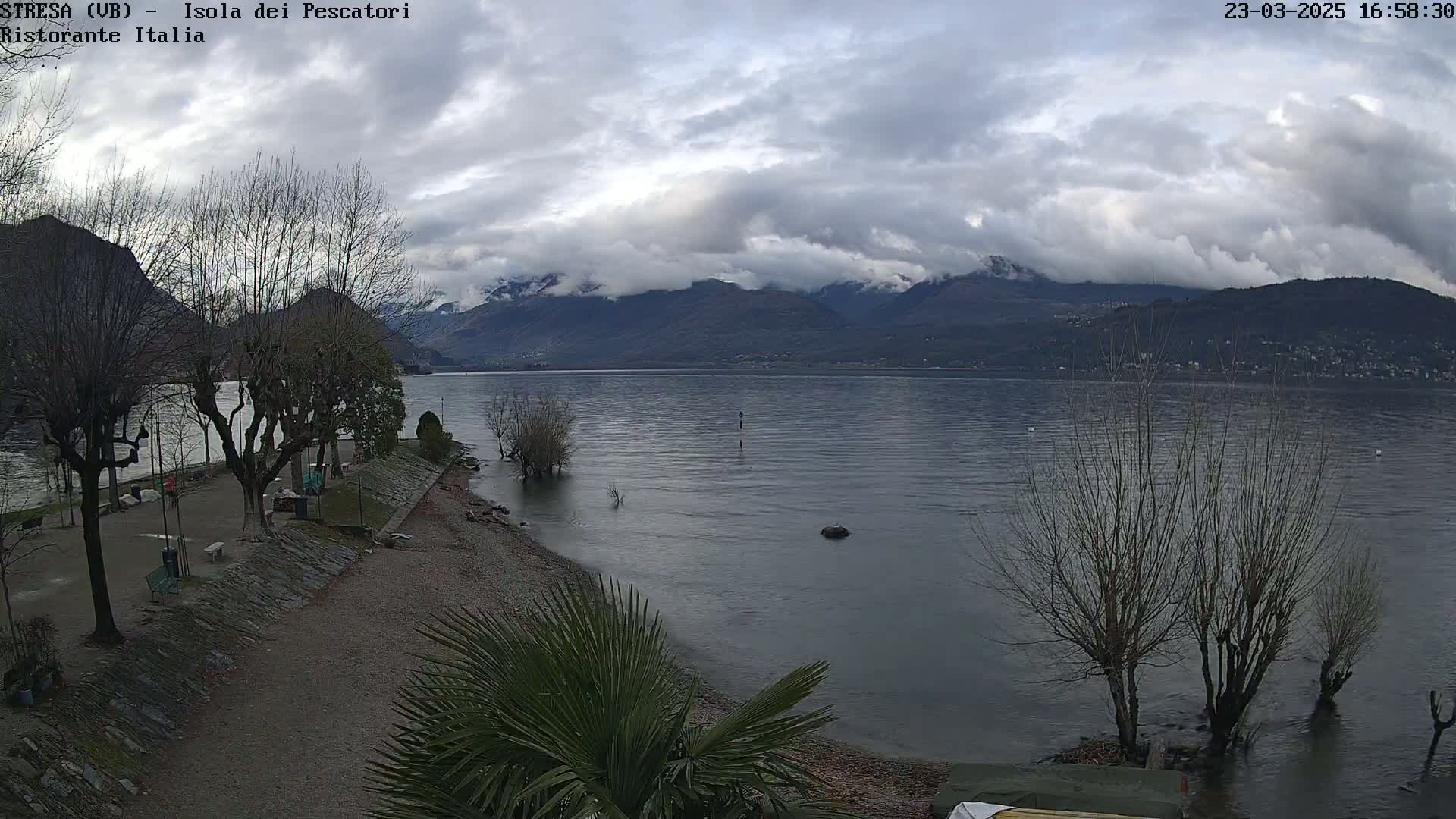 A mostly cloudy day reveals a calm lake with mountains in the background, seen from a lakeside park with leafless trees and a sandy beach.