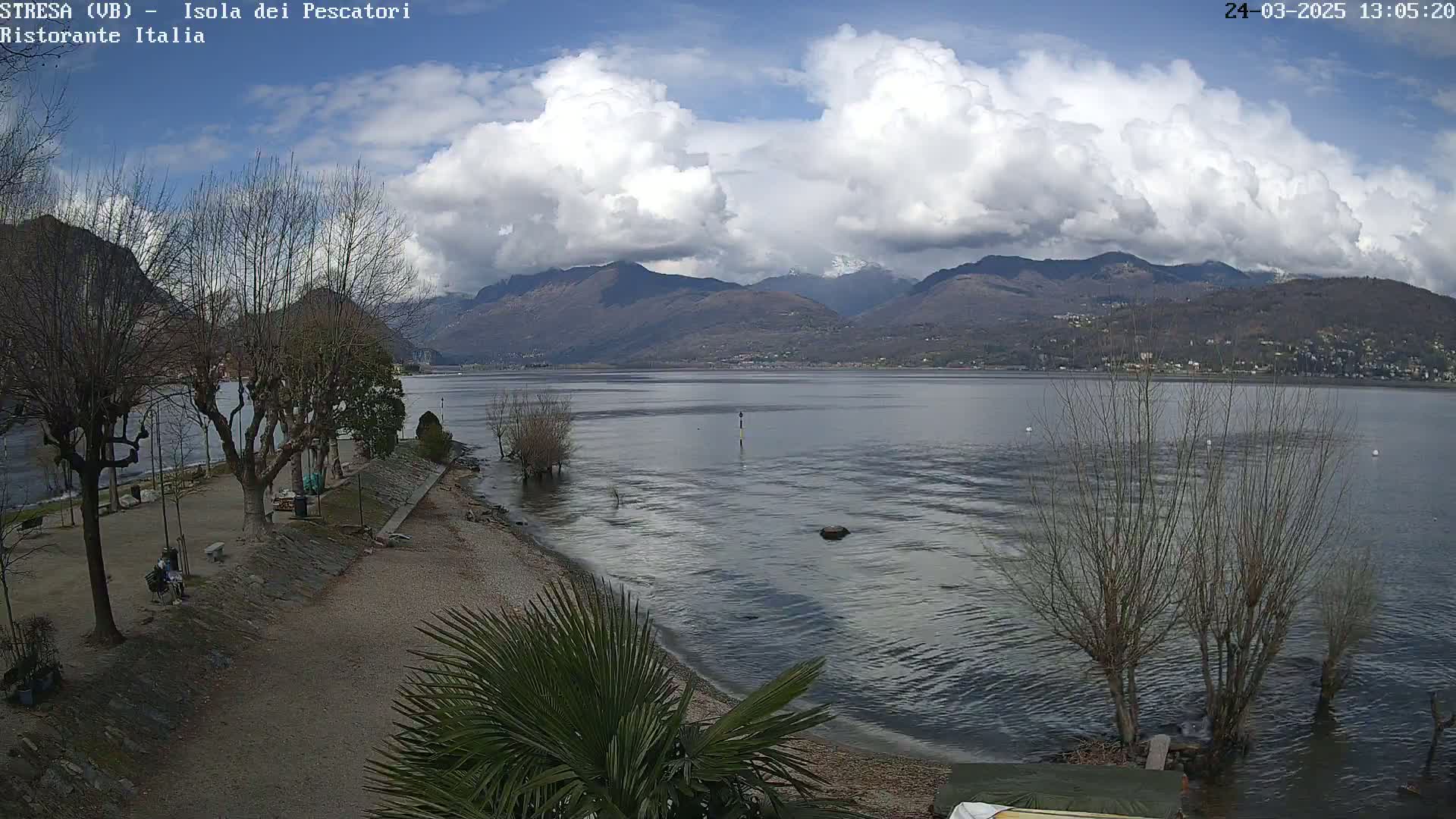 A partly cloudy day shows a calm lake with mountains in the background, a shoreline with bare trees and a sandy beach, and a person sitting on a bench.