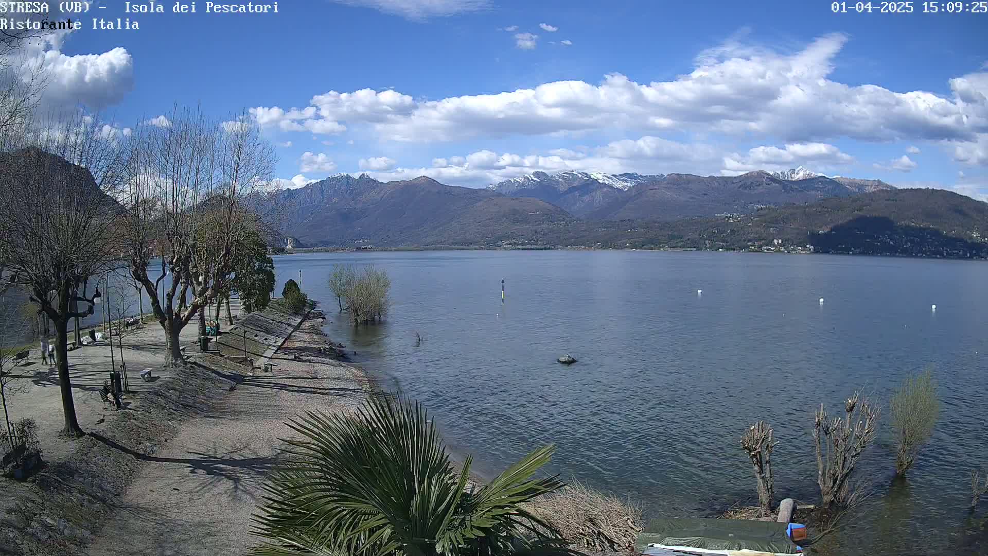 A partly sunny day reveals a calm lake with snow-capped mountains in the background, viewed from a lakeside park with bare trees and a sandy shore.