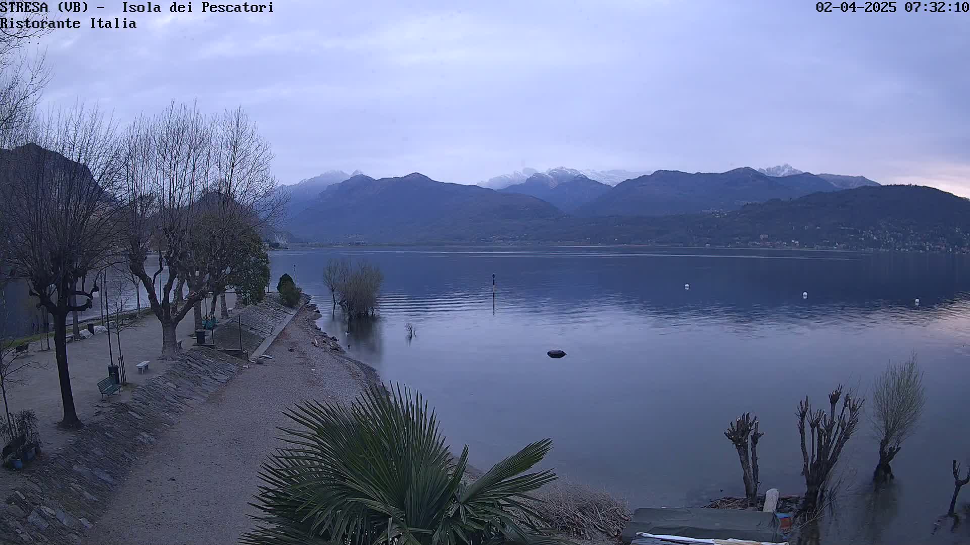 A calm lake under an overcast sky is nestled beside a shoreline with bare trees and a sandy beach, backed by snow-capped mountains in the distance.