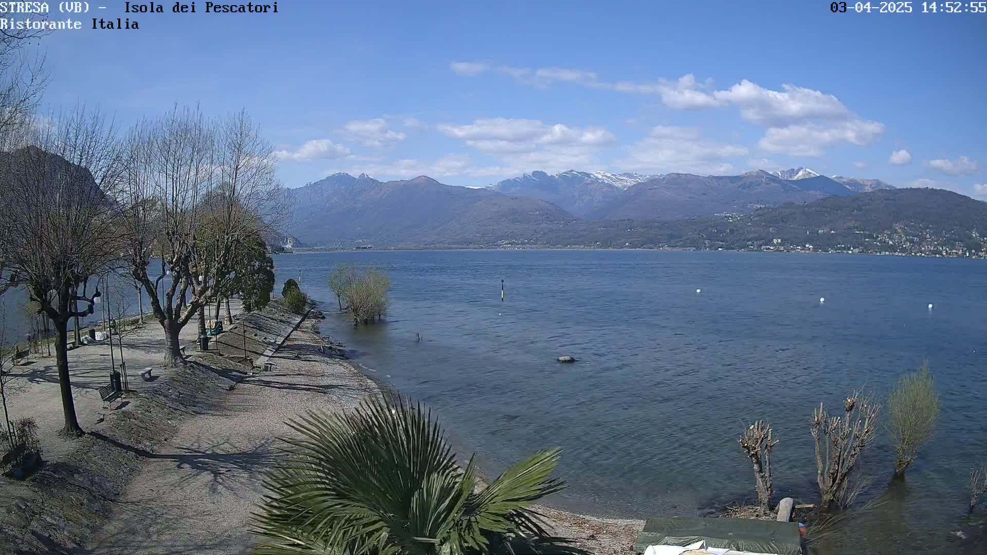 A mostly sunny day reveals a lakeside view with leafless trees, a sandy shoreline, snow-capped mountains in the distance, and a calm body of water.