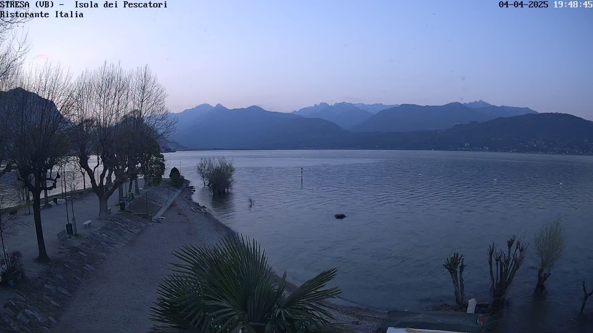 A tranquil lake scene at dusk shows bare trees lining a shore with a small beach, under a clear sky, mountains in the distance.
