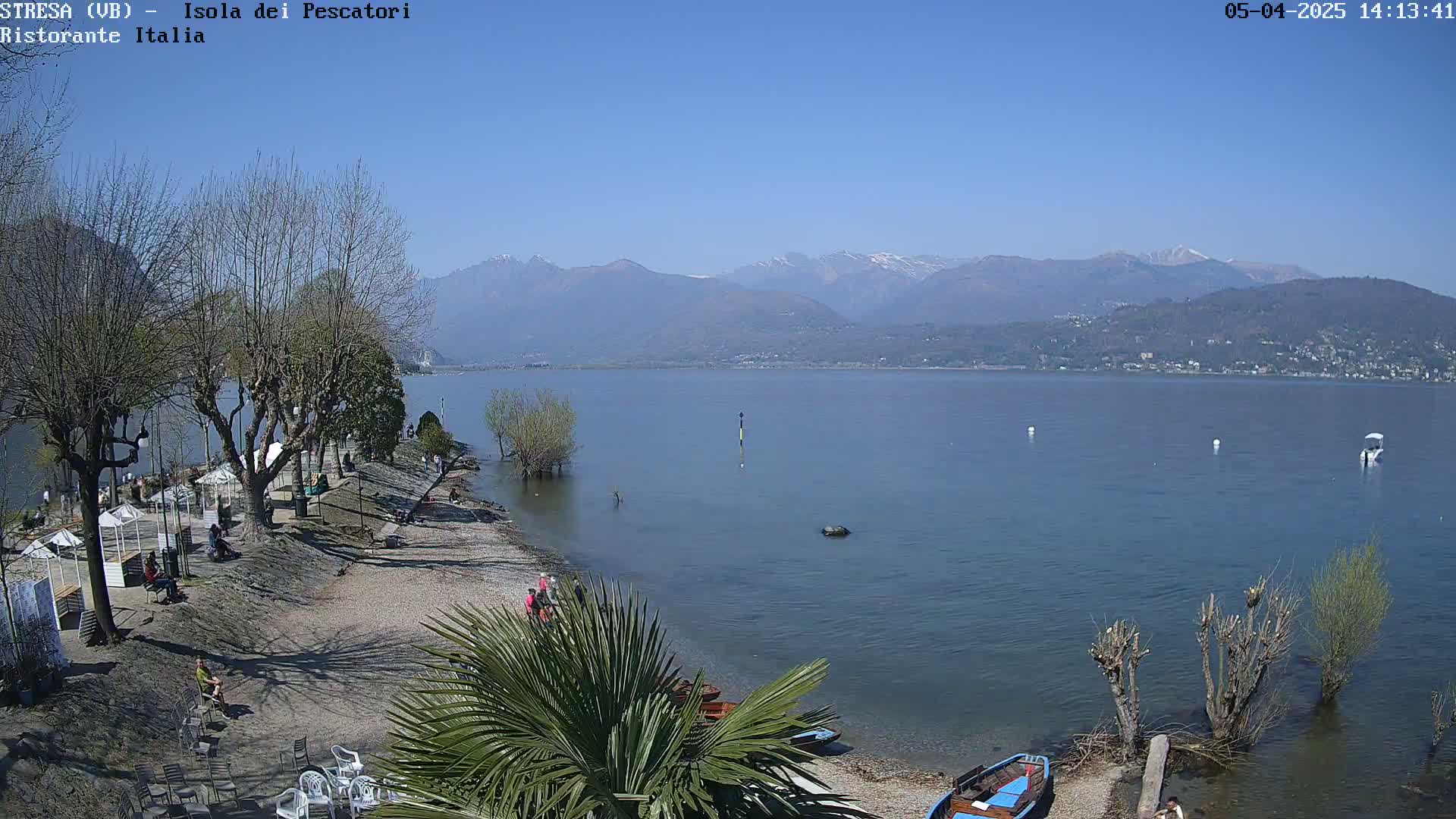 A sunny day overlooking a calm lake with mountains in the background, a rocky shoreline with trees and several small boats, and a few people scattered along the shore.