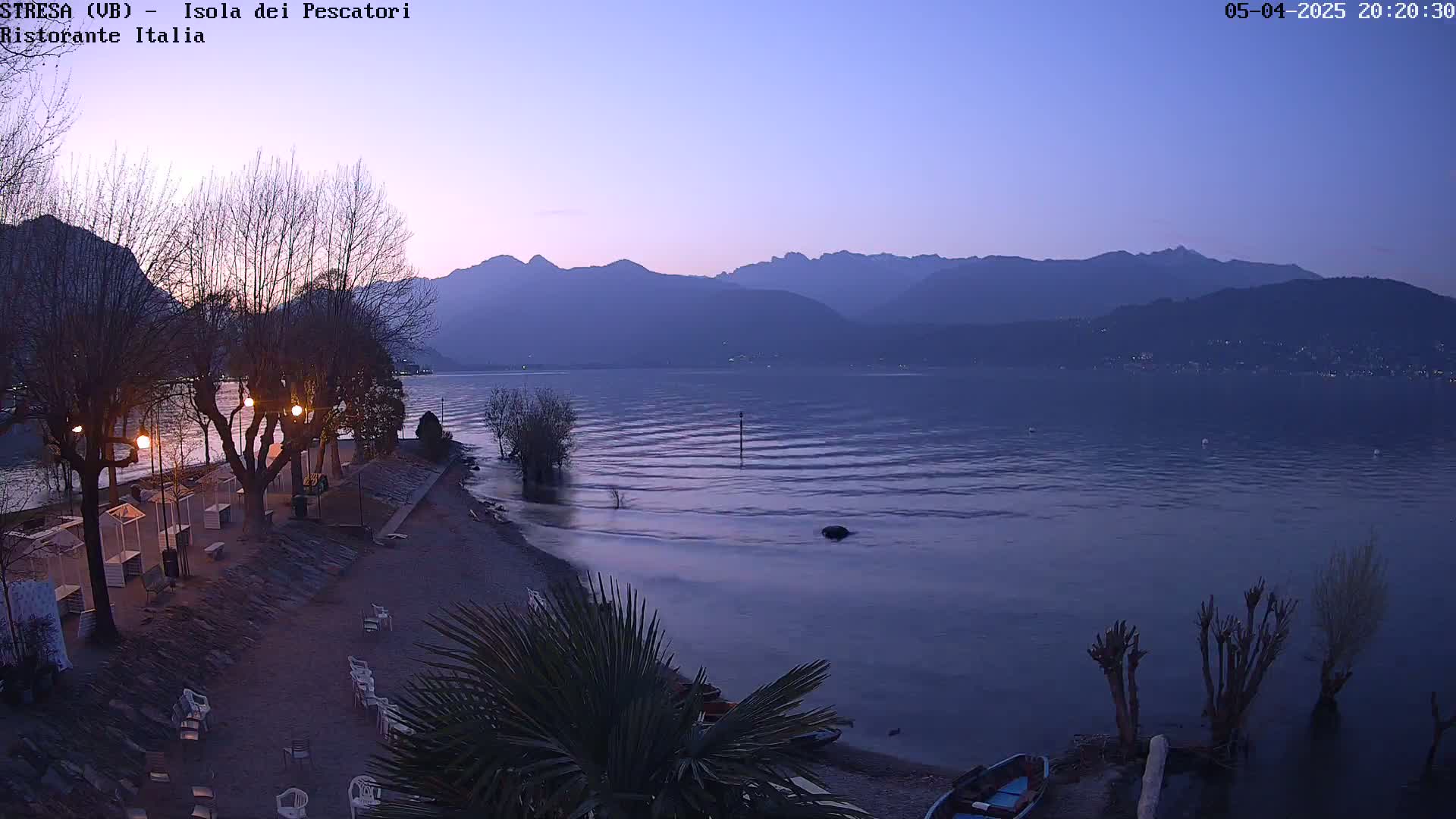 A calm lake at twilight is bordered by a shoreline with bare trees, lights, and a few small boats, backed by a mountain range.