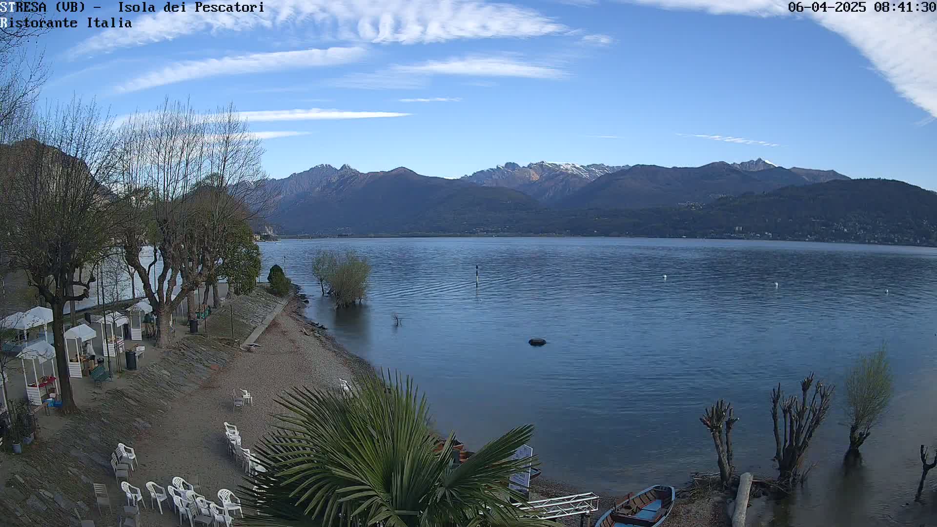 A clear, sunny day shows a calm lake with mountains in the background, a sandy beach with several trees and some small boats at the water's edge.