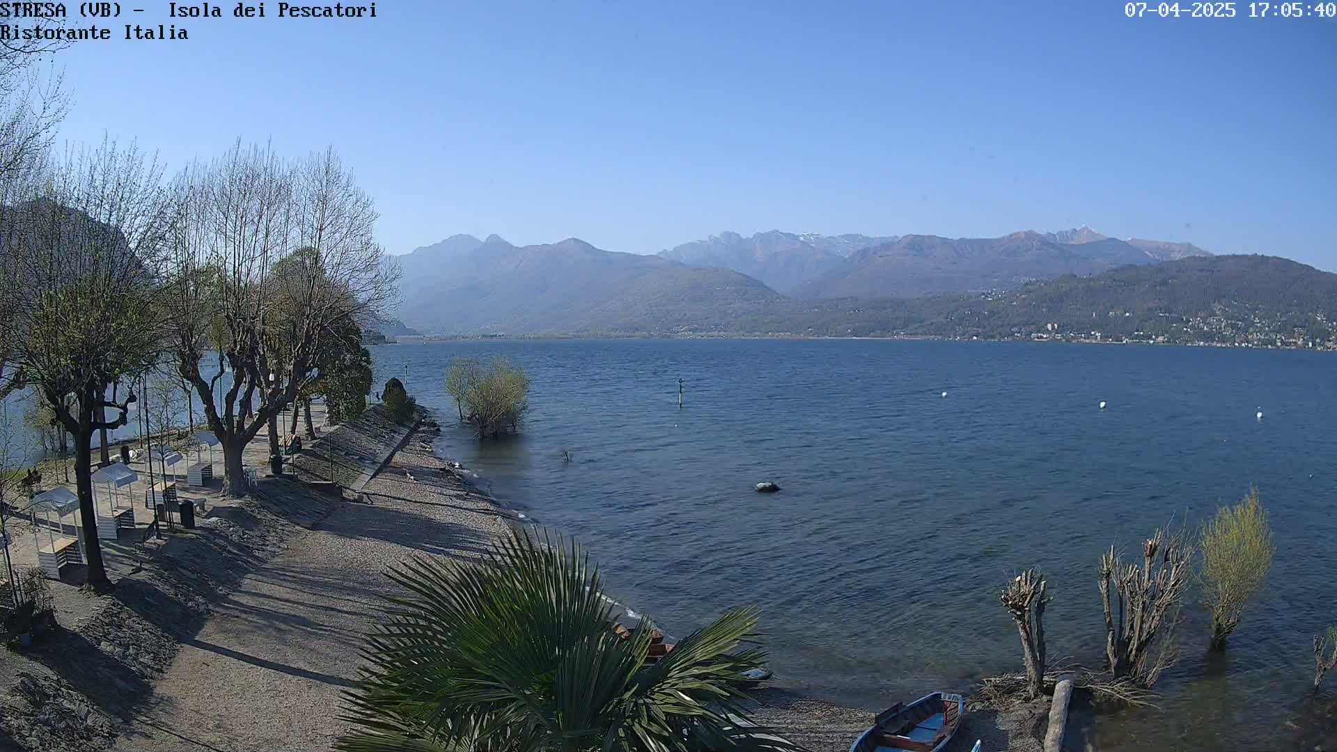 A tranquil lake scene under a clear blue sky shows a sandy shore with leafless trees, a few small boats, and distant mountains.
