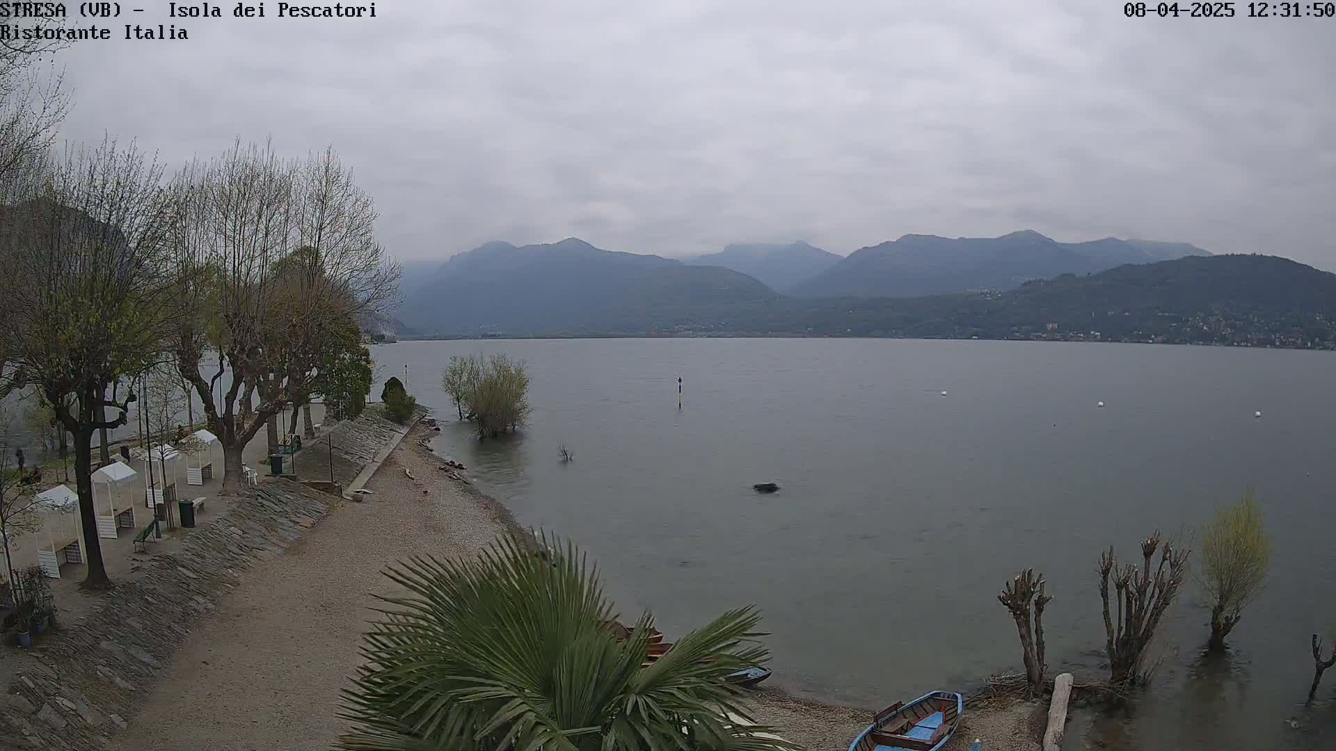 A view of a calm lake under an overcast sky, with mountains in the background, a pebble beach in the foreground, and several trees and small boats along the shoreline.