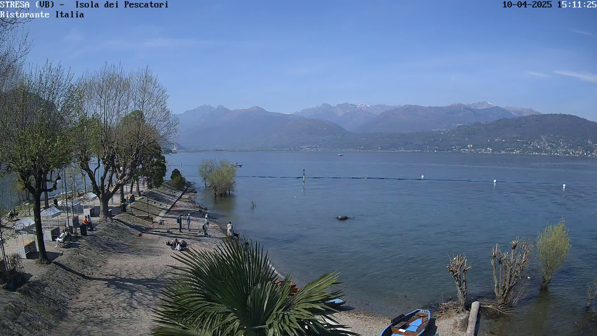 A calm lake under a clear blue sky is viewed from a shoreline with several people, bare trees, and a palm tree, with mountains in the background.