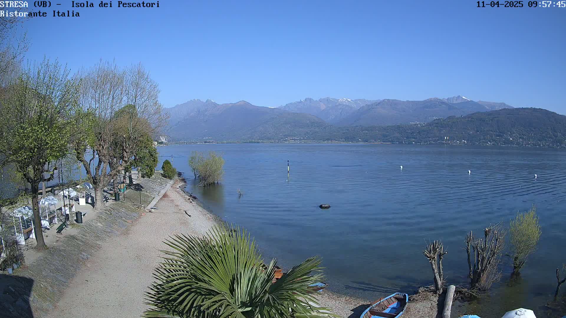 A clear, sunny day reveals a calm lake with mountains in the background, a sandy shore with trees and a few boats, and some beach umbrellas.