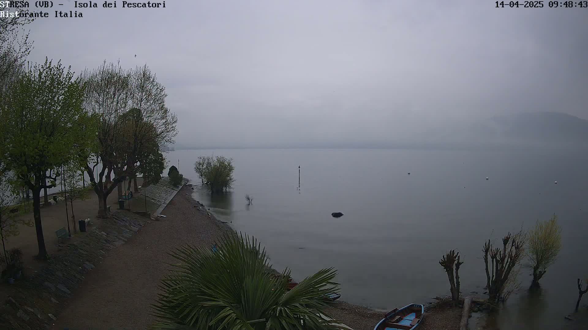 A hazy, overcast day shows a calm lake with a few boats pulled ashore near a rocky beach, lined with trees and a paved walkway.