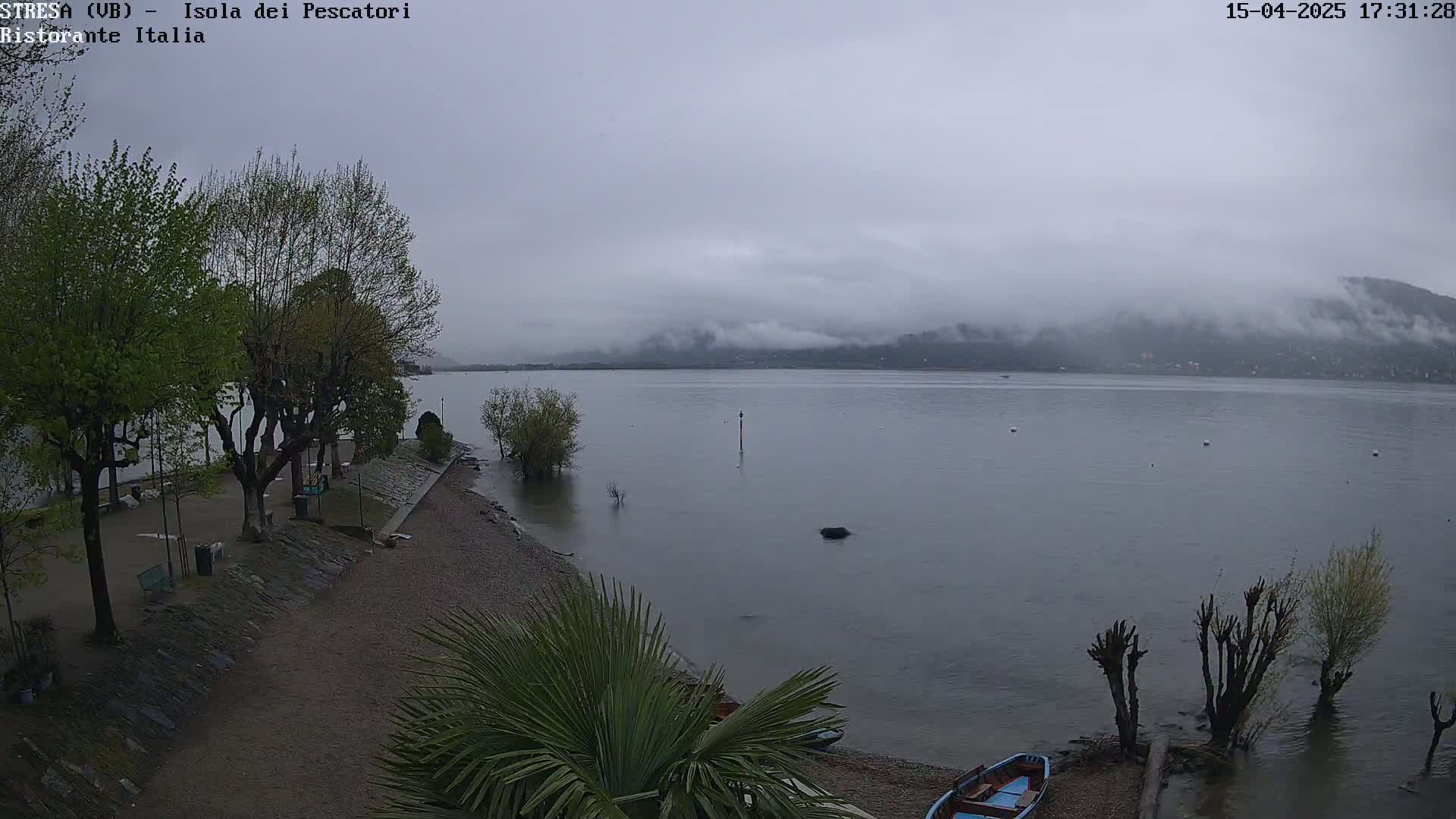 A calm lake under an overcast sky, partially shrouded in fog, is seen from a lakeside park with trees and a small boat on the shore.