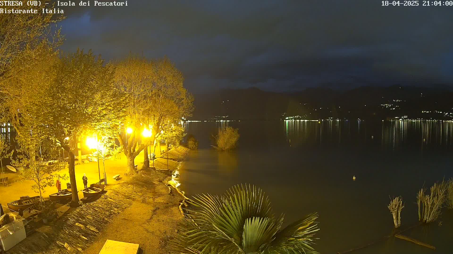 A nighttime view of a calm lake reflecting lights from a town across the water, with illuminated trees and several small boats on a shore in the foreground.