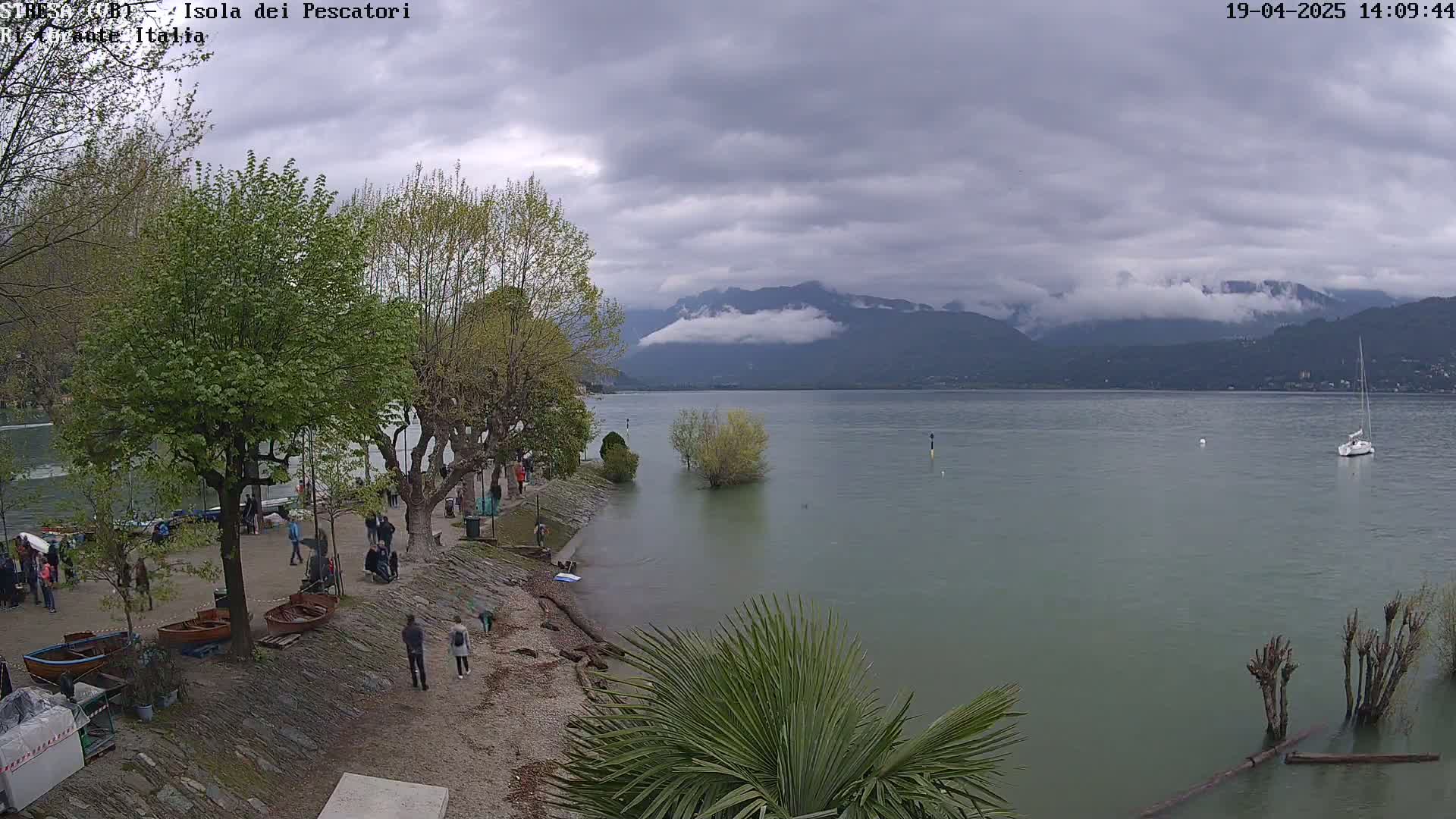 A lake with a partly cloudy sky, high water levels flooding part of the shoreline near trees and people, and mountains in the background.