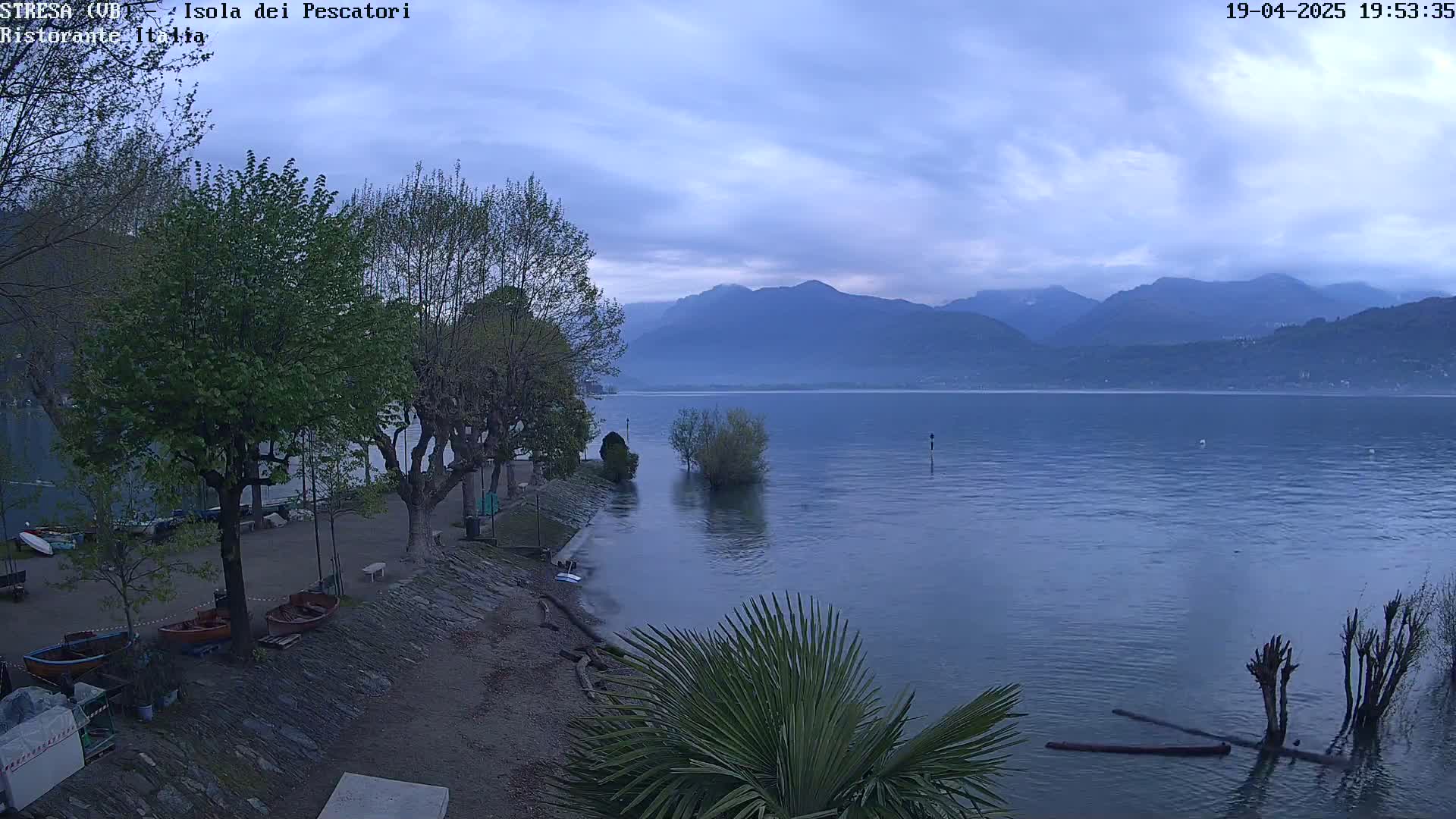A calm lake at dusk is seen from a lakeside vantage point, with several trees and small boats visible near a low stone wall, under an overcast sky and mountains in the background.