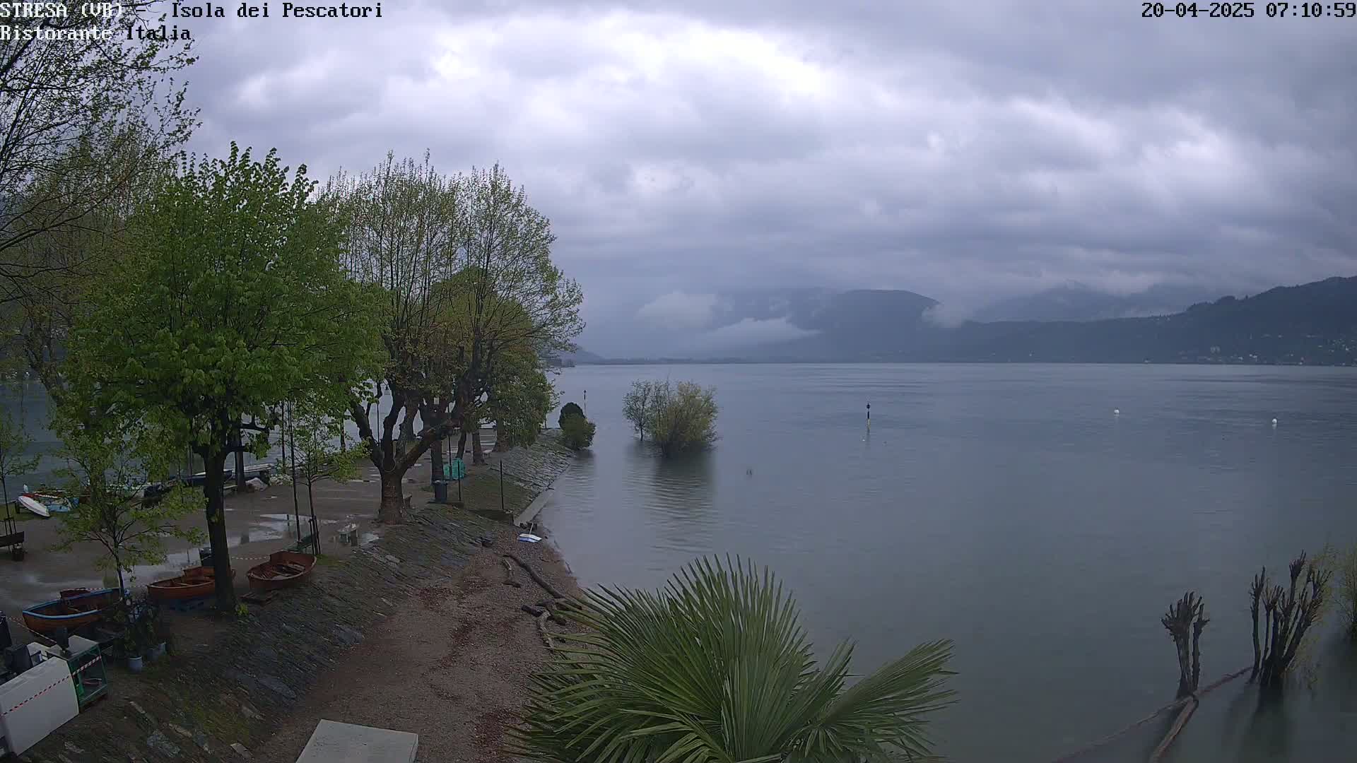 A cloudy, overcast day shows a calm lake with several small boats pulled ashore near leafy trees and a partially submerged shoreline.