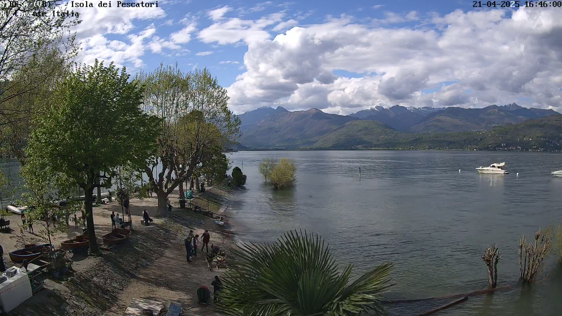 A partly cloudy day shows a lake with a shoreline featuring people, trees, and small boats, with mountains in the background.