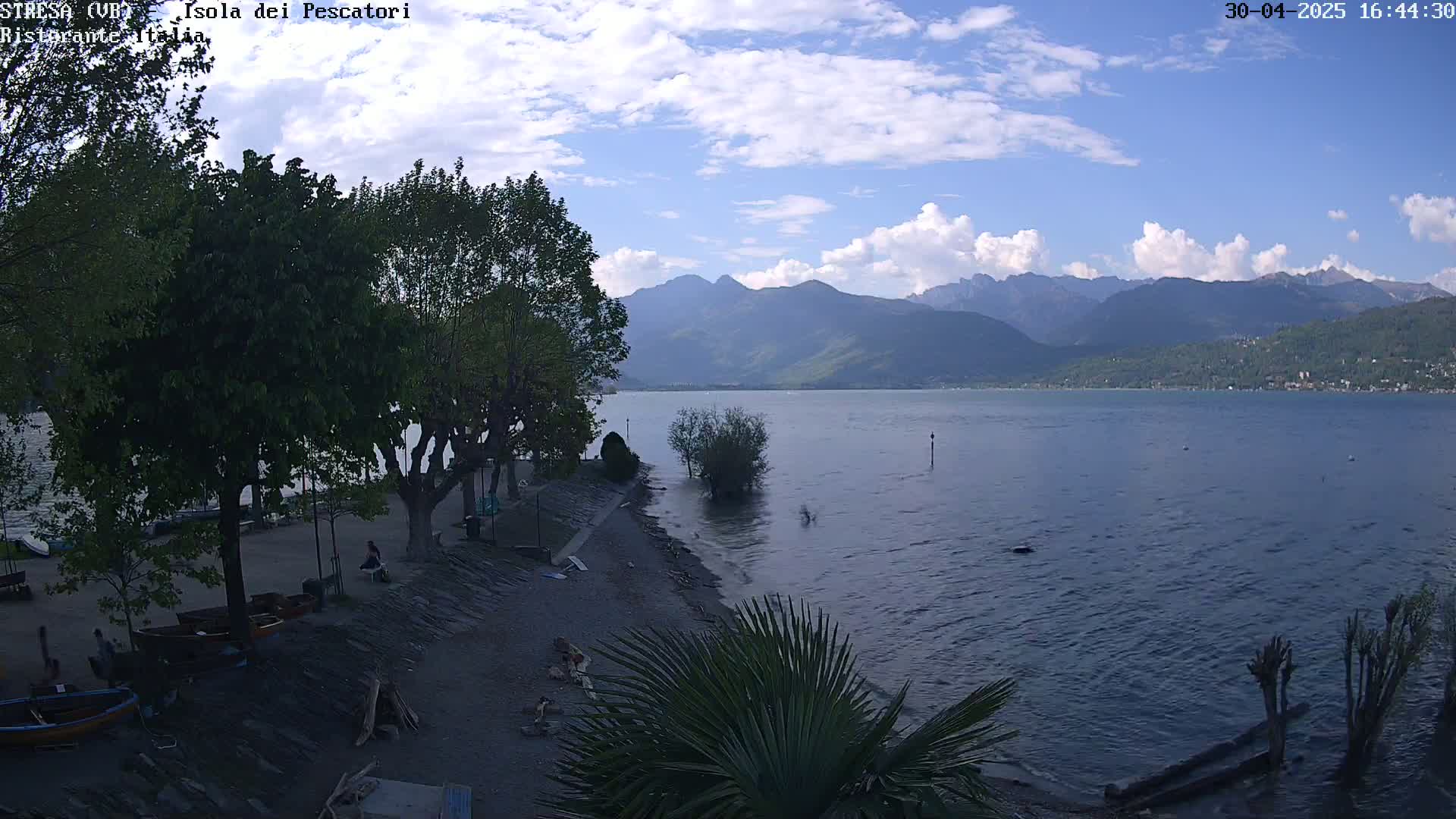 A partly cloudy day shows a lake shore with trees, boats, and people, overlooking a calm lake and distant mountains.