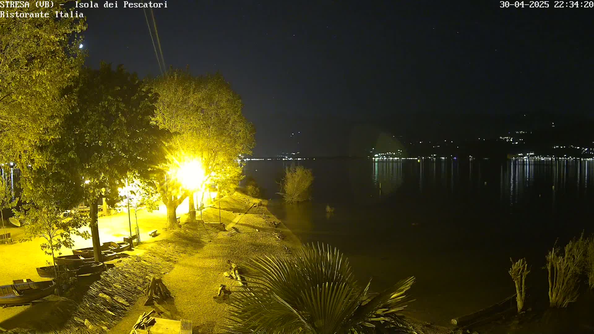 A nighttime lakeside scene shows a calm body of water reflecting lights from a distant town, with a shoreline illuminated by artificial lights near several small boats and trees.